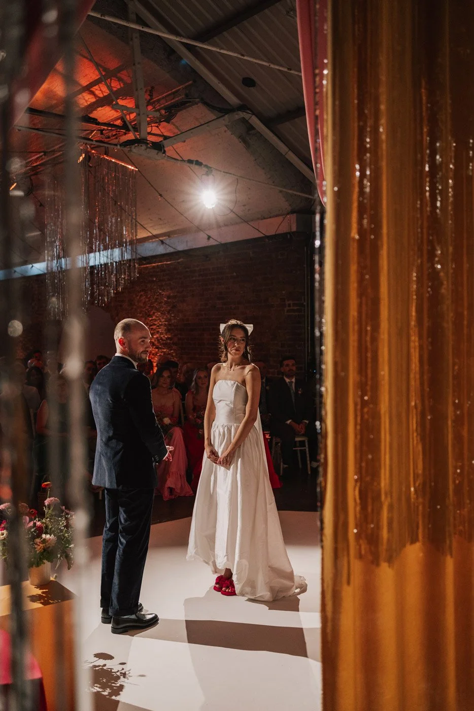 A bride and groom standing during a wedding ceremony, with the bride in a white dress and the groom in a dark suit, in a dimly lit venue with exposed brick walls and a spotlight overhead.