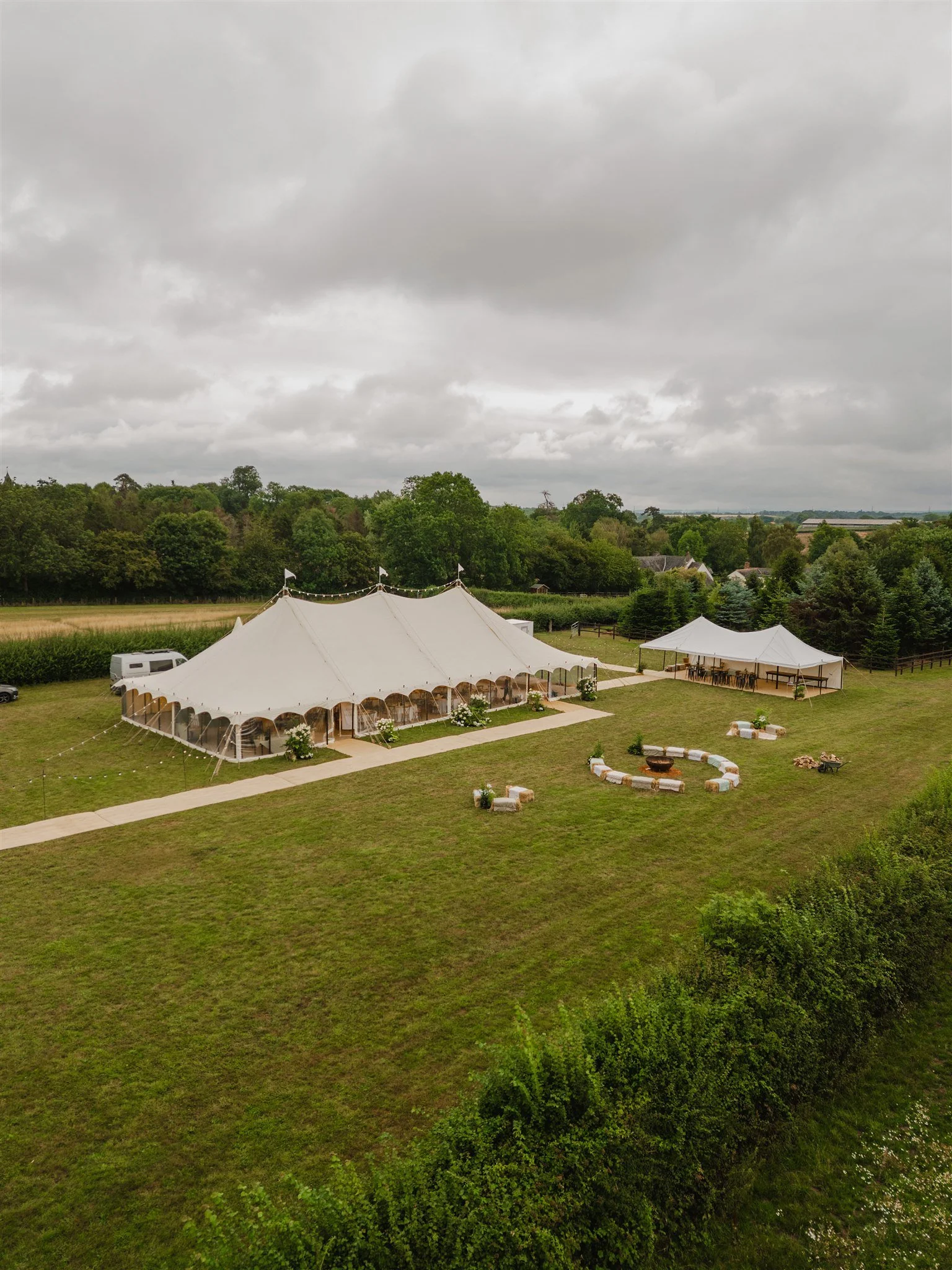 Large white event tent set up on a grassy field with smaller tents nearby, surrounded by greenery and trees, under cloudy skies.