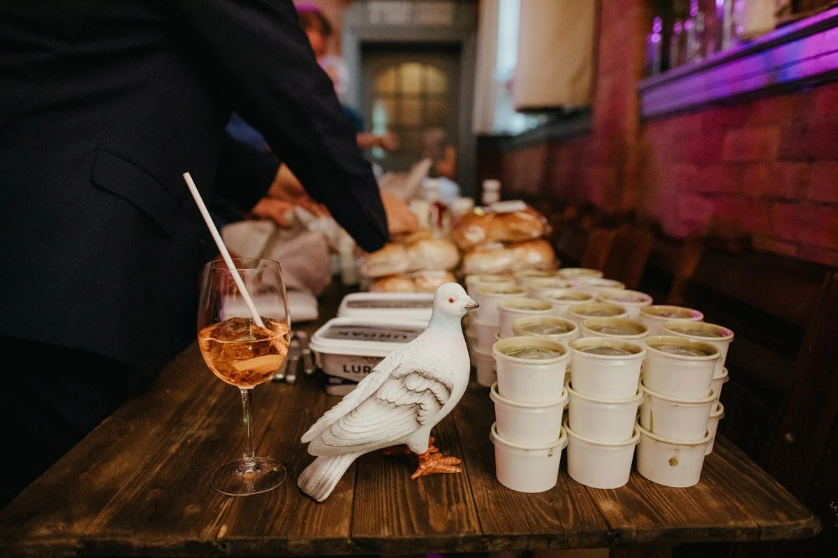A wooden table with a white decorative dove, a glass of pink-colored drink with a straw, and several white cups arranged in rows. In the background, people are serving food, and there are stacks of bread or sandwiches.