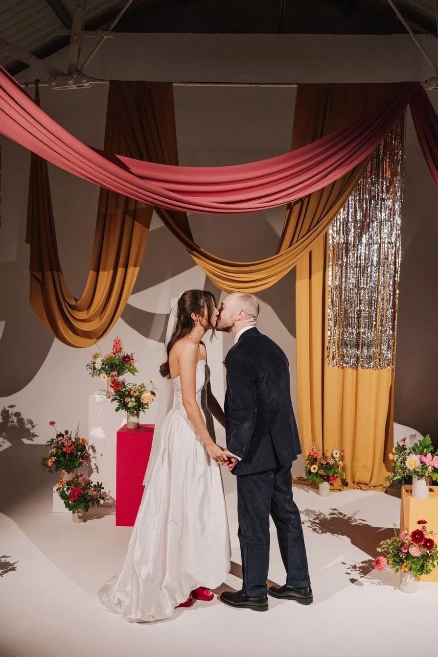 Bride and groom holding hands and kissing during their wedding ceremony, decorated with colorful flowers and draped curtains.