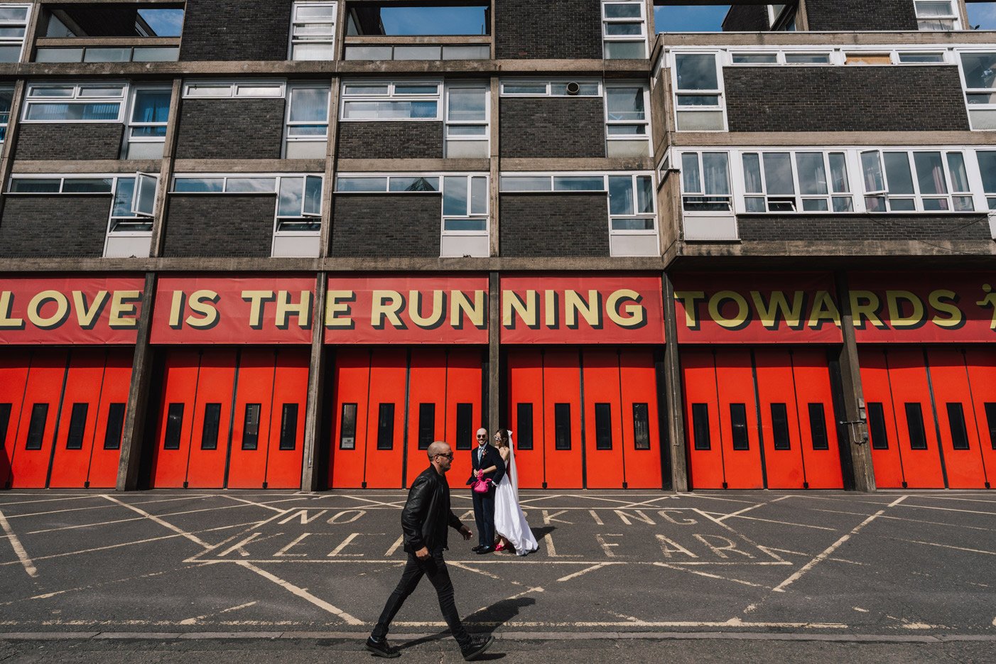 A man walking in front of a building with a large red and yellow sign that reads 'LOVE IS THE RUNNING TOWARDS' and a couple, with the woman in a white dress and the man in a dark suit, standing near a donation bin.