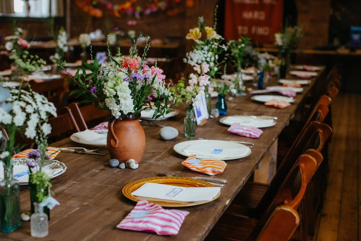 Long wooden table decorated with flowers in various vases, set for a meal with plates, napkins, and utensils, in a rustic indoor setting.