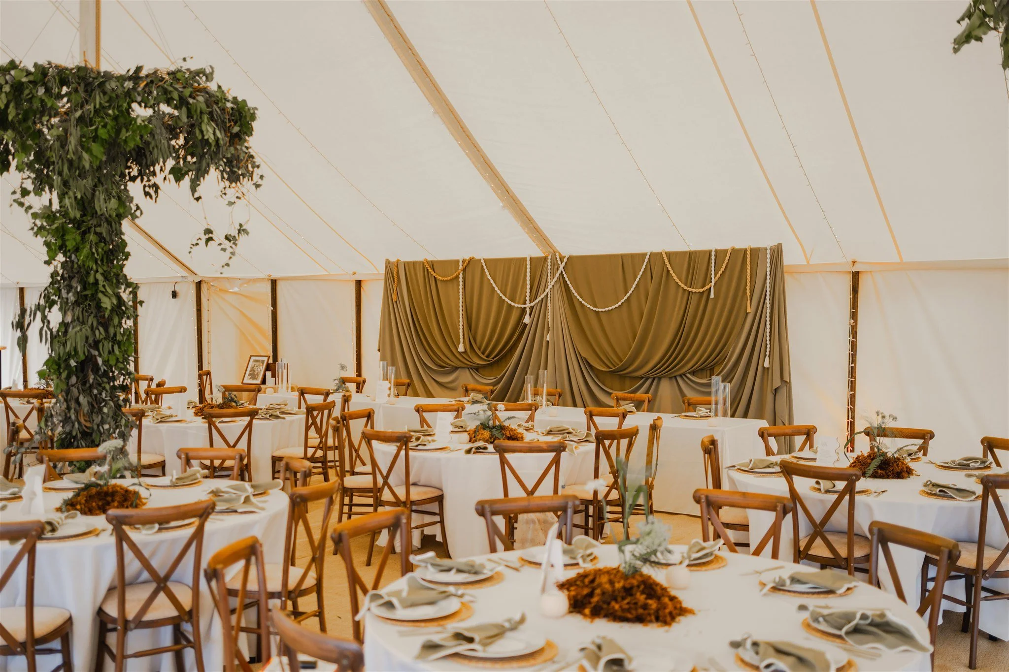 Decorated event tent with round tables covered in white tablecloths, set with napkins, plates, and centerpieces, and a backdrop with draped fabric and garlands.