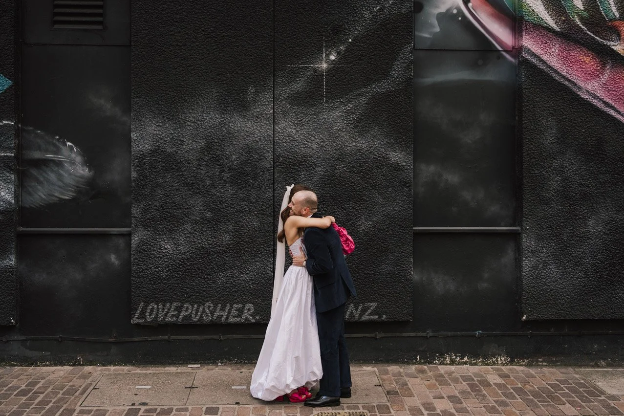 A couple embraces in front of a large black mural on a city street. The woman is wearing a white dress, and the man is in a suit. The mural features a whale and what appears to be fireworks or stars, with the text "LOVE PUSHER" and possibly a URL or name below.