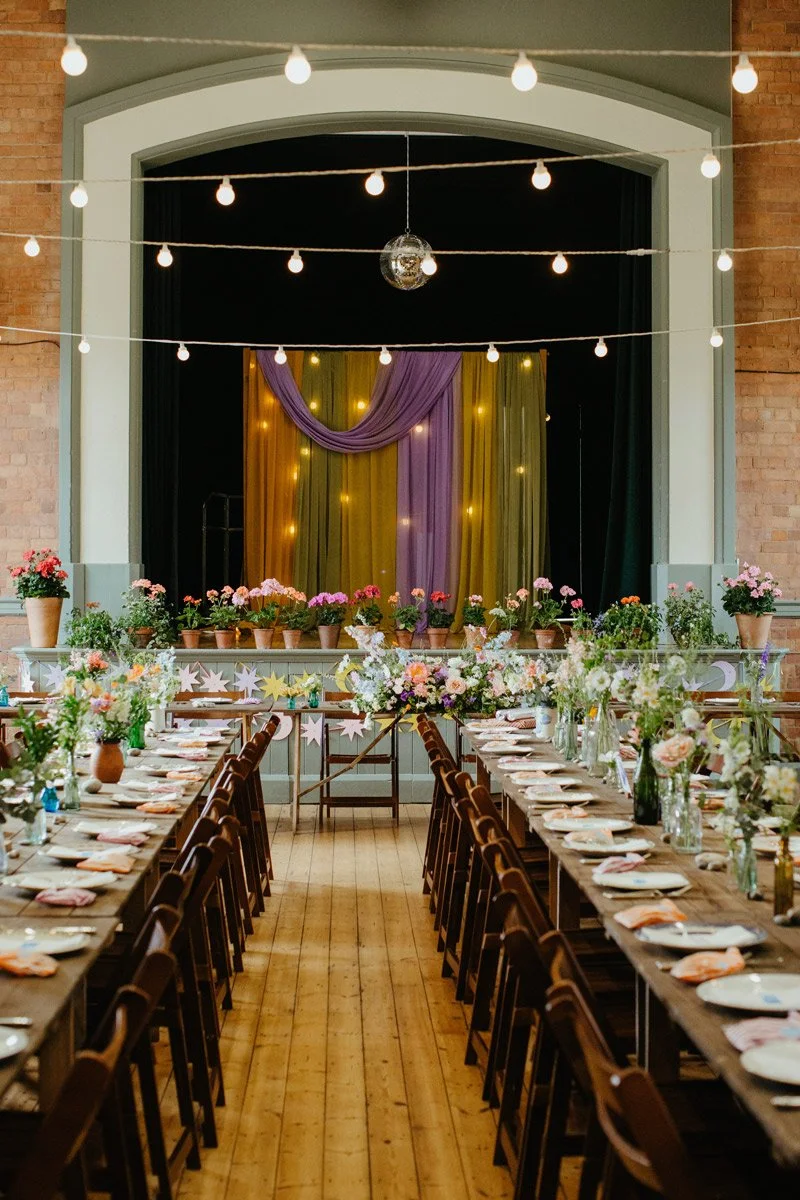 Decorated indoor dining area with string lights, a stage with purple and yellow curtains, floral arrangements on tables, and potted plants along the stage.