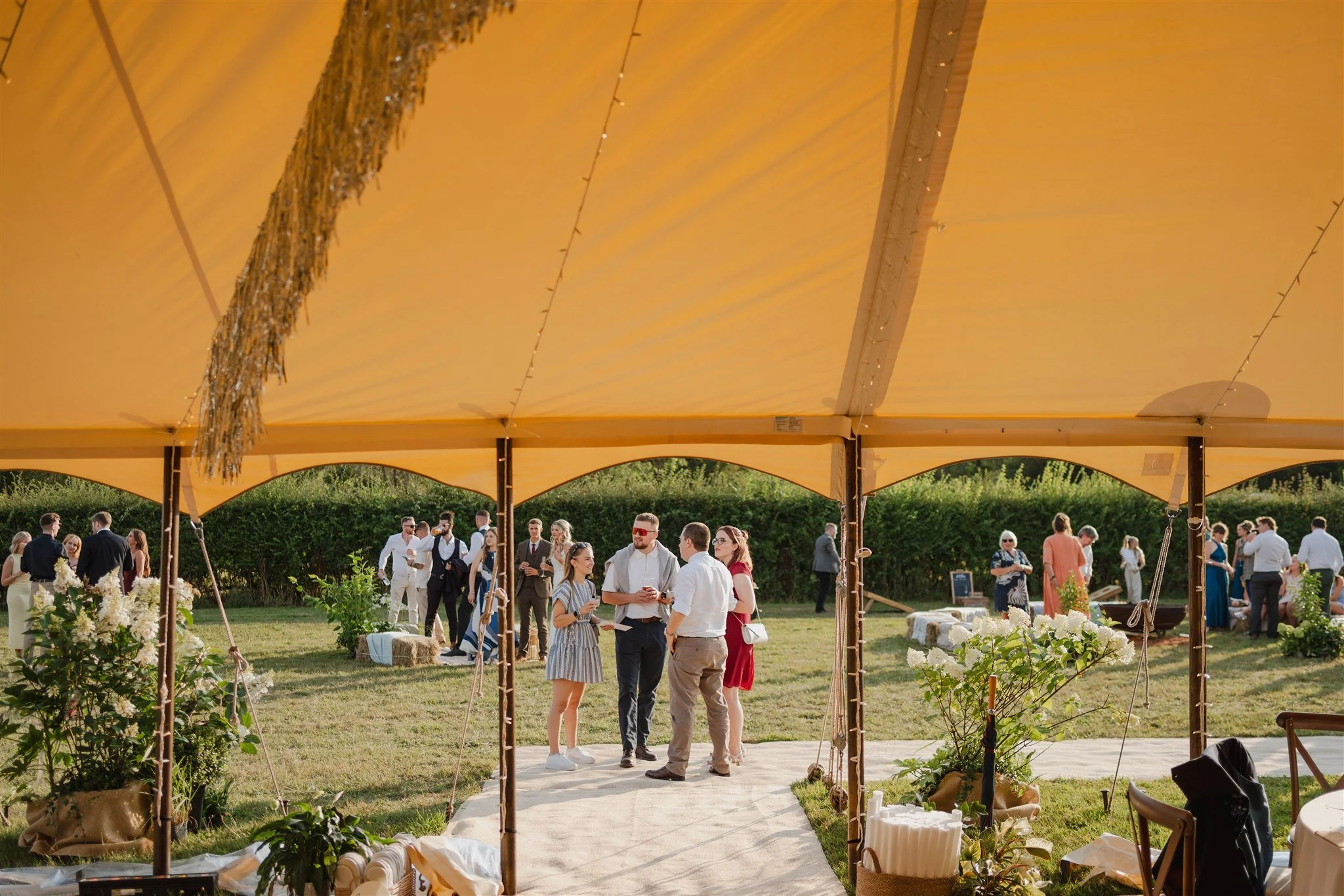 Guests socializing at an outdoor celebration under a yellow canopy with greenery and sunlight.