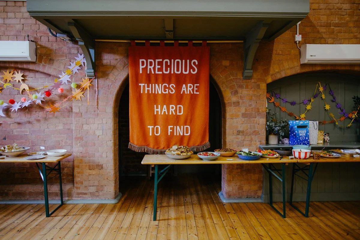 Decorative table with bowls of food, colorful hanging decorations, a large orange banner that says "Precious Things Are Hard to Find," and a wrapped gift box in a brick-walled room.