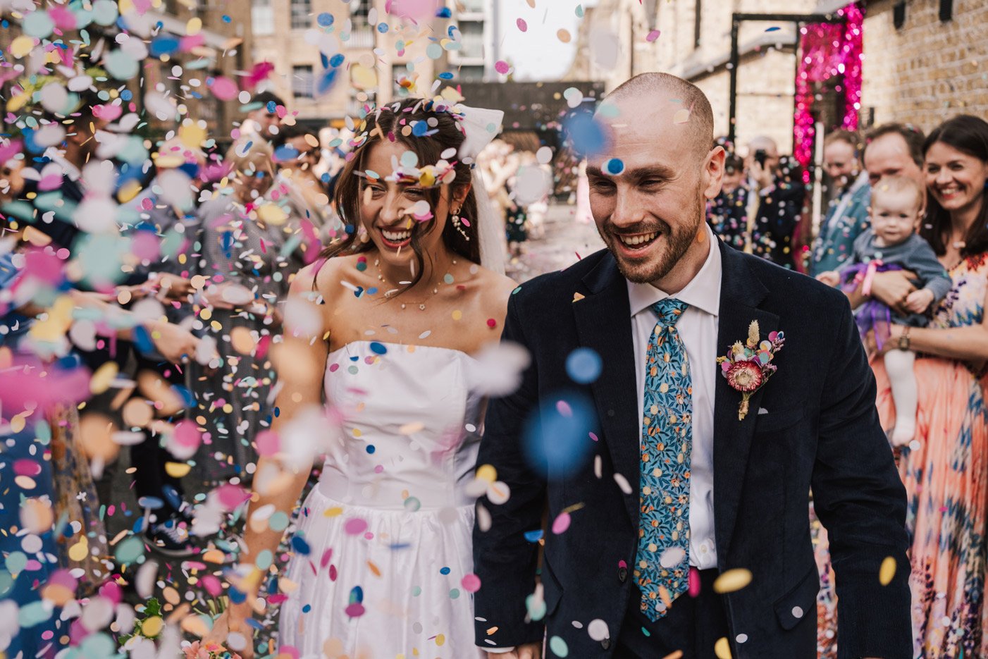 A joyful bride and groom celebrate at their wedding amid colorful confetti, with friends and family in the background.