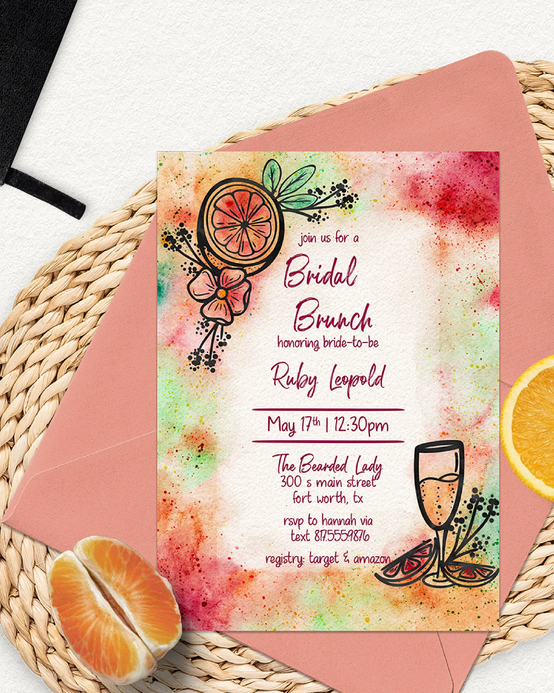 Colorful watercolor-style bridal brunch invitation with a slice of orange, floral and leaf illustrations, and champagne glass illustration, laid on a paper and woven placemat, with orange slices nearby.