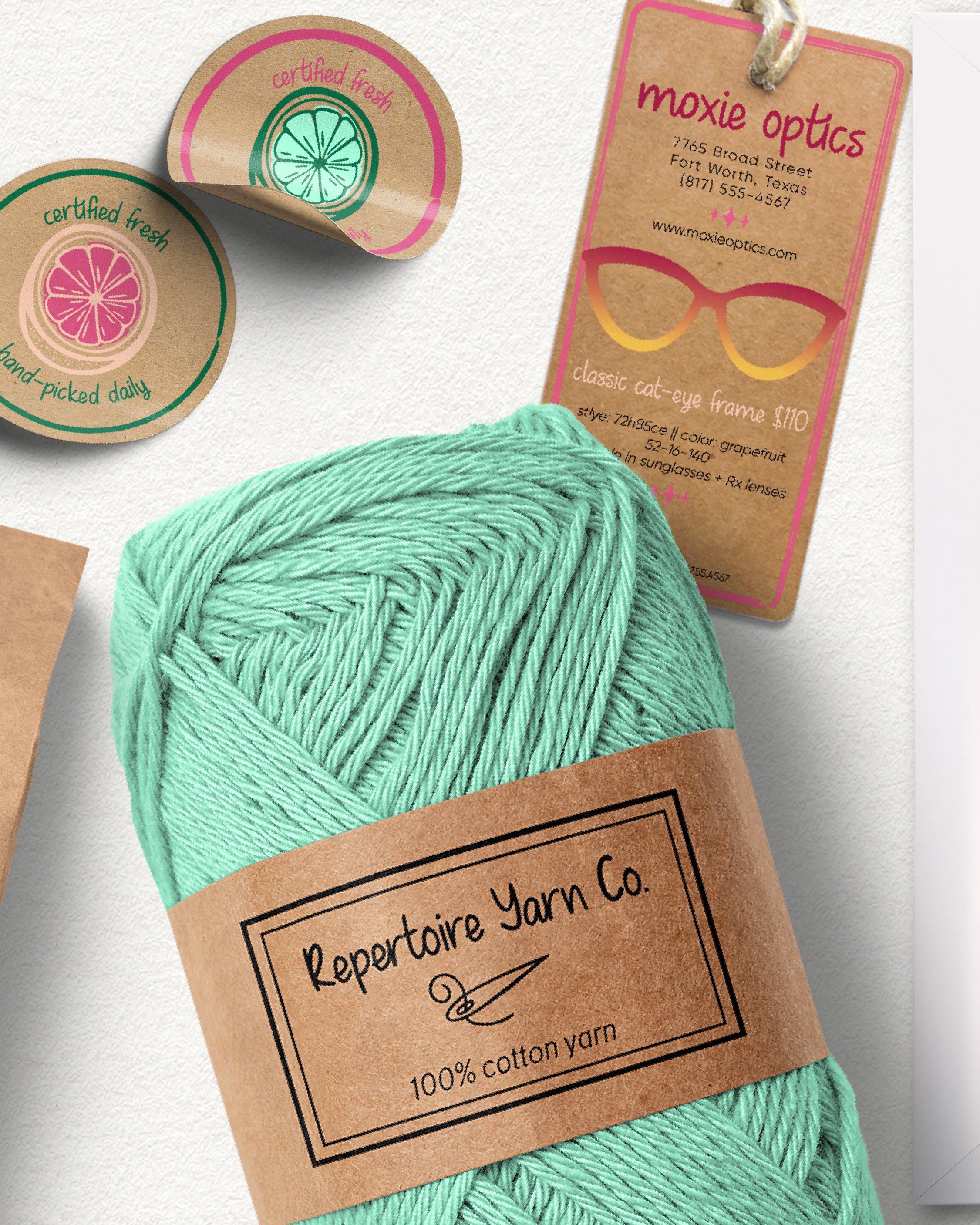 Close-up of mint green yarn with brown craft paper labels featuring colorful citrus slice illustrations and text, including details for a craft yarn brand and a business card for Moxie Optics.