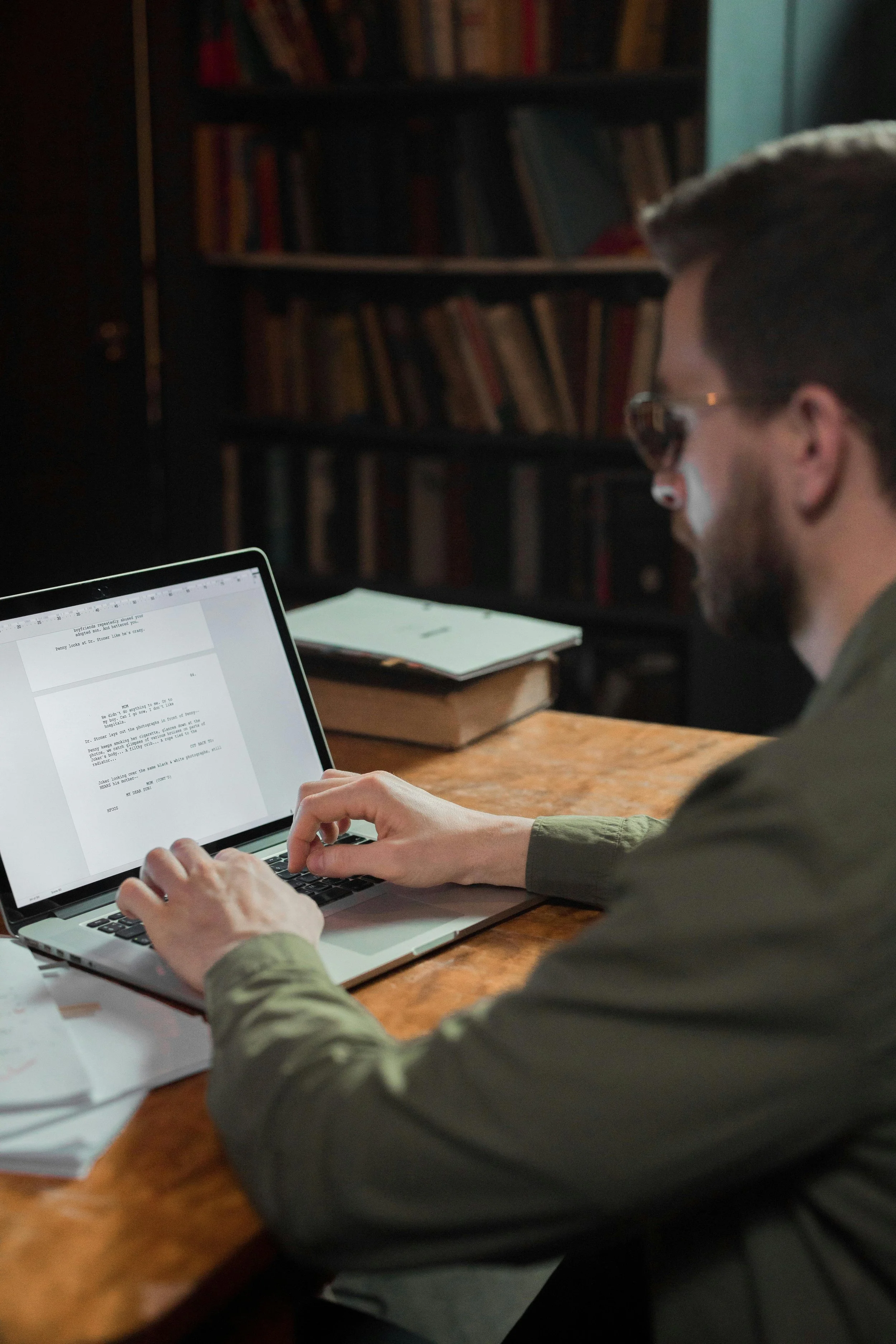 A person wearing sunglasses and a green shirt is working on a laptop at a wooden desk in a room with bookshelves in the background.