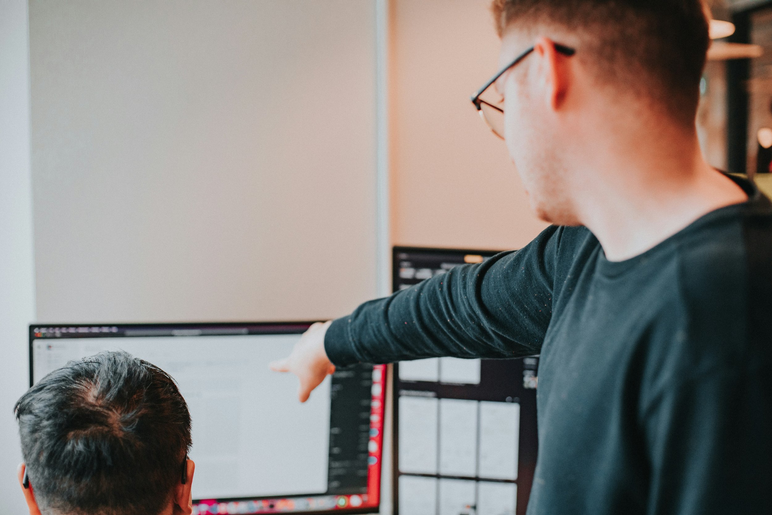 A man wearing glasses points at a computer screen with another person sitting in front of it, both working on a project together.