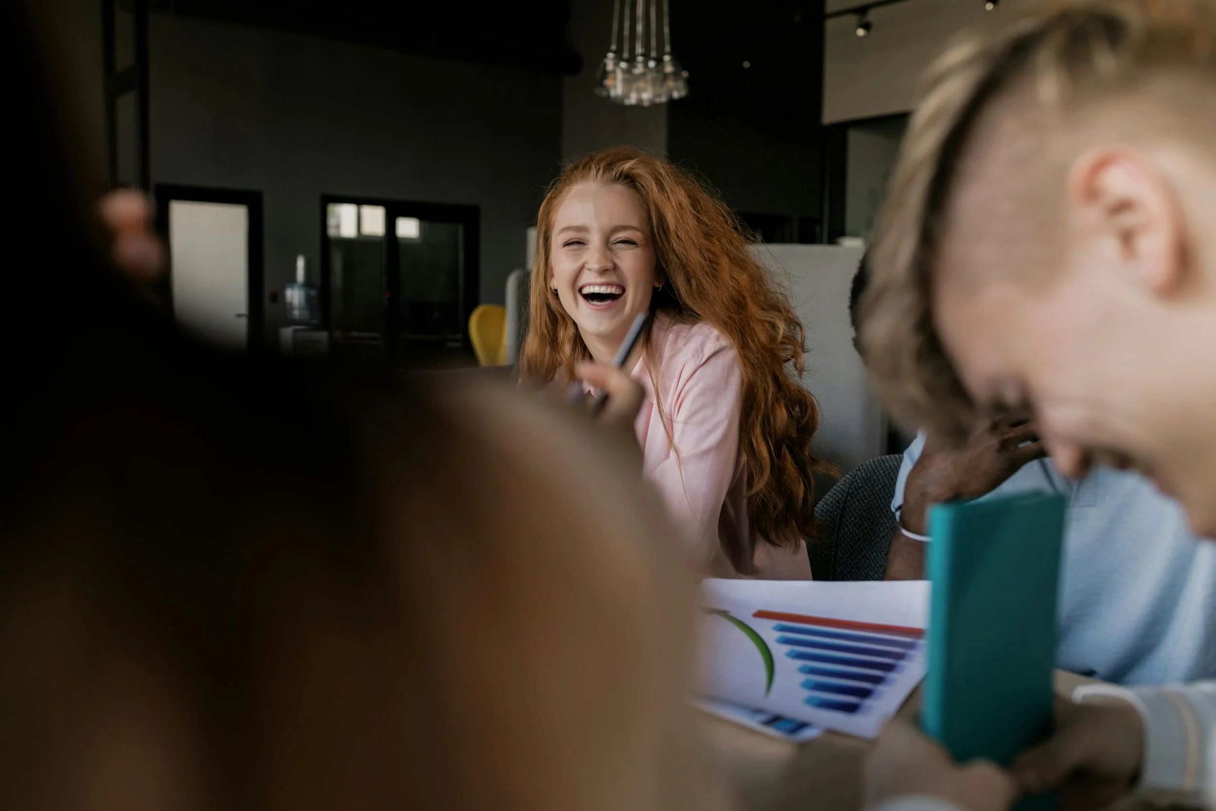 A woman with red hair and a pink blazer laughing during a meeting with colleagues in a modern office.