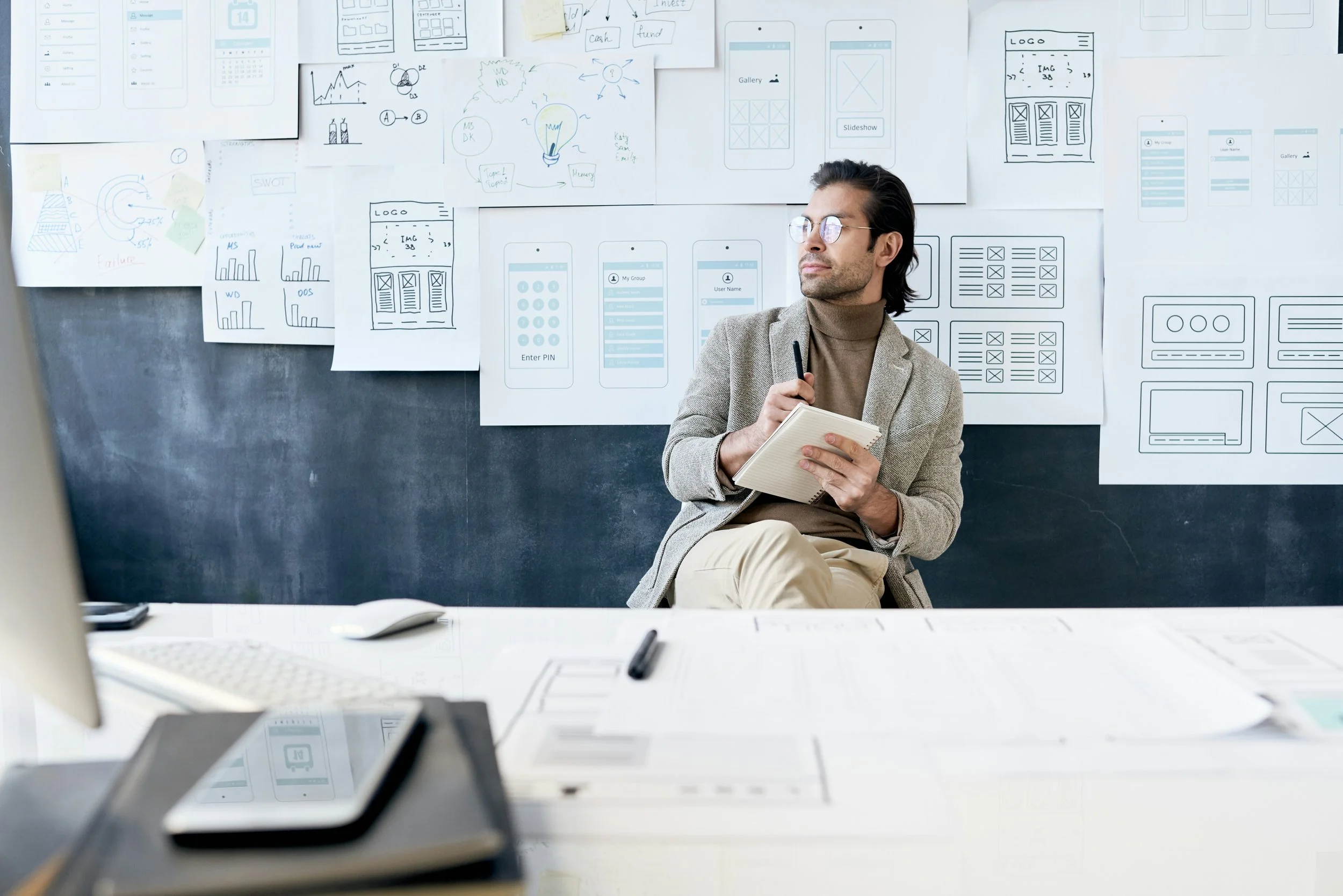 A man sitting in a meeting room with creative sketches and wireframes pinned to the wall behind him, holding a notebook and pen, appearing to be deep in thought.