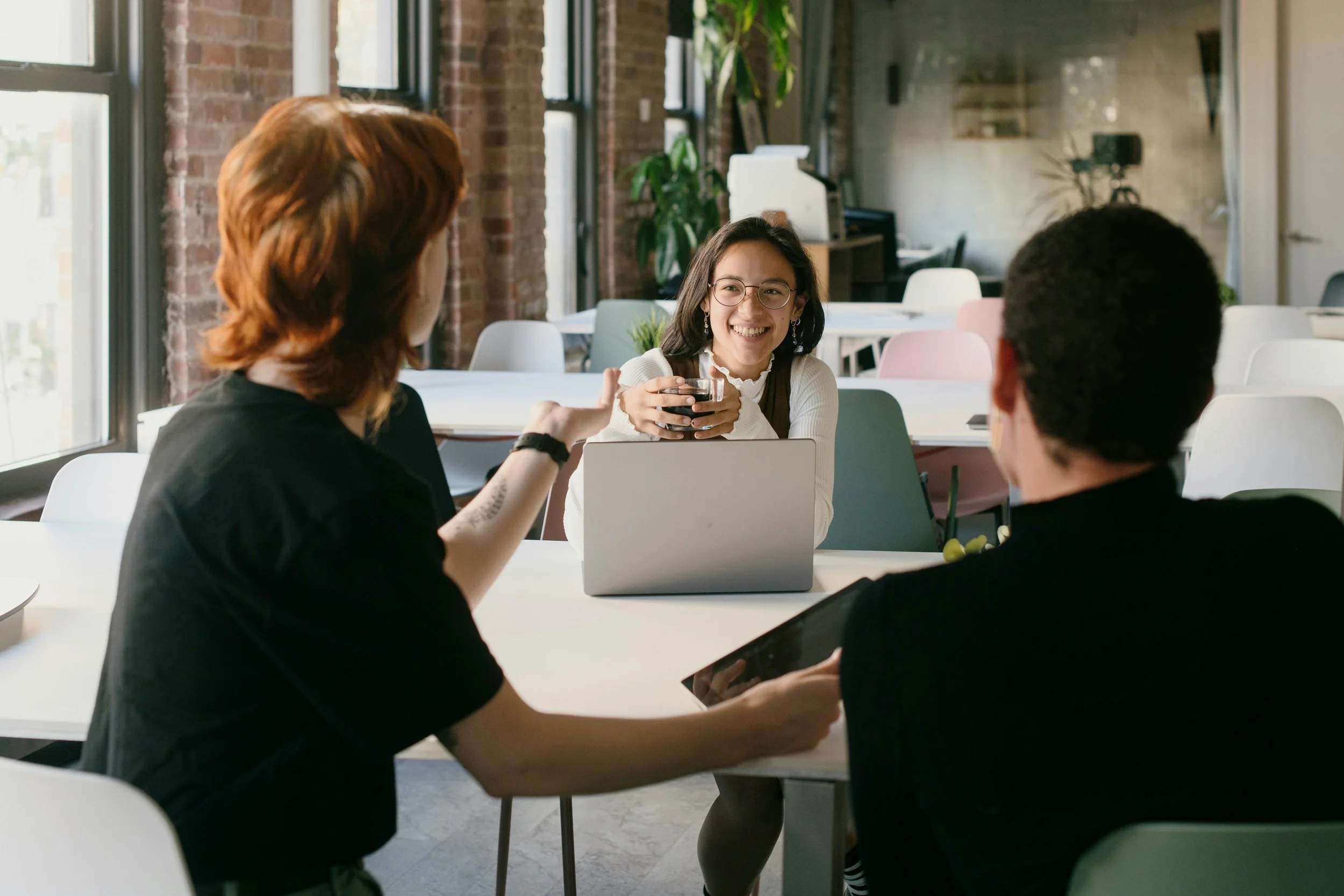 Three people having a conversation at a table in a modern office space, with the woman in the center smiling and holding a coffee mug.