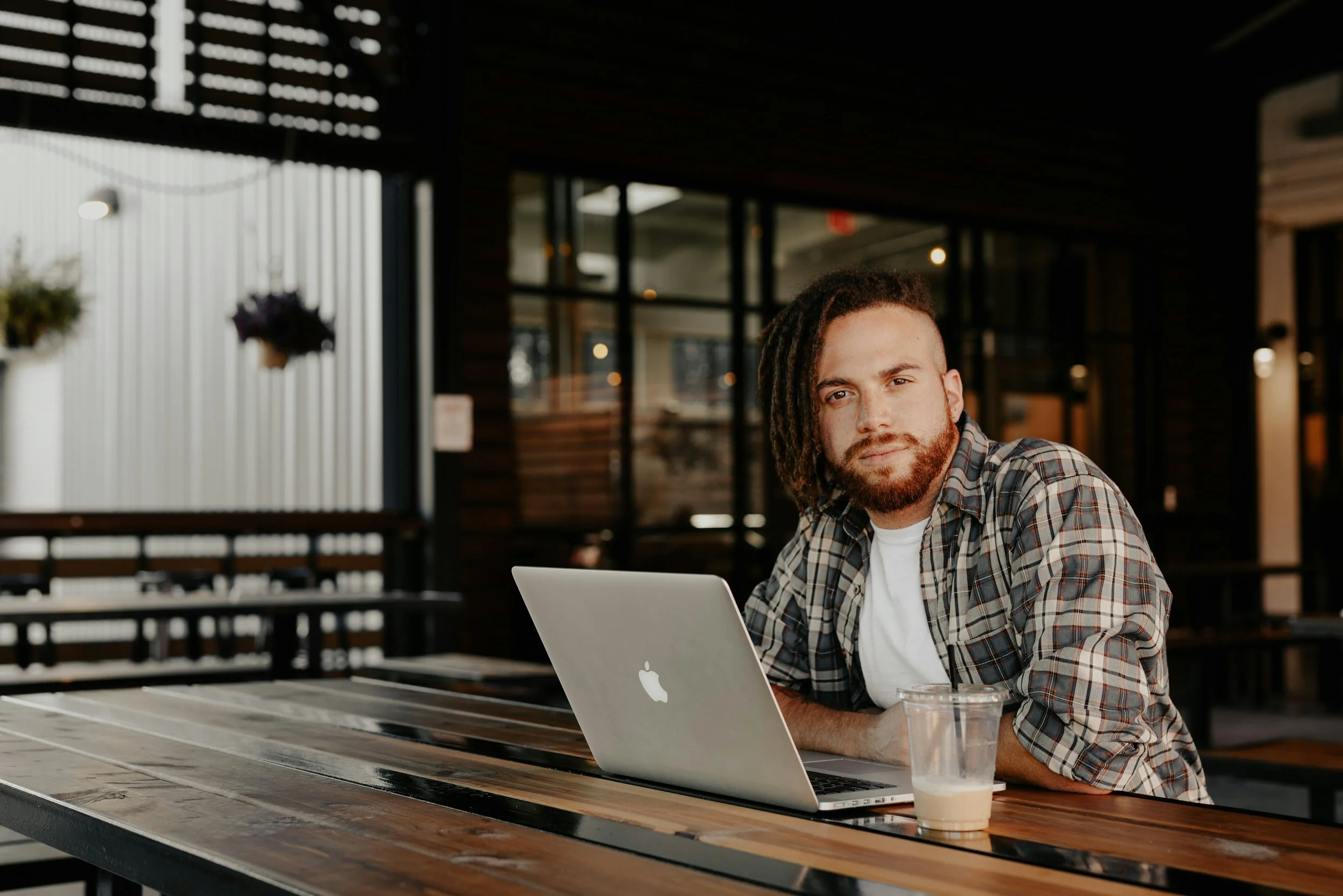 A man with dreadlocks and a beard sitting at a wooden table with a MacBook, inside a modern cafe with glass and wood decor, with a latte on the table.
