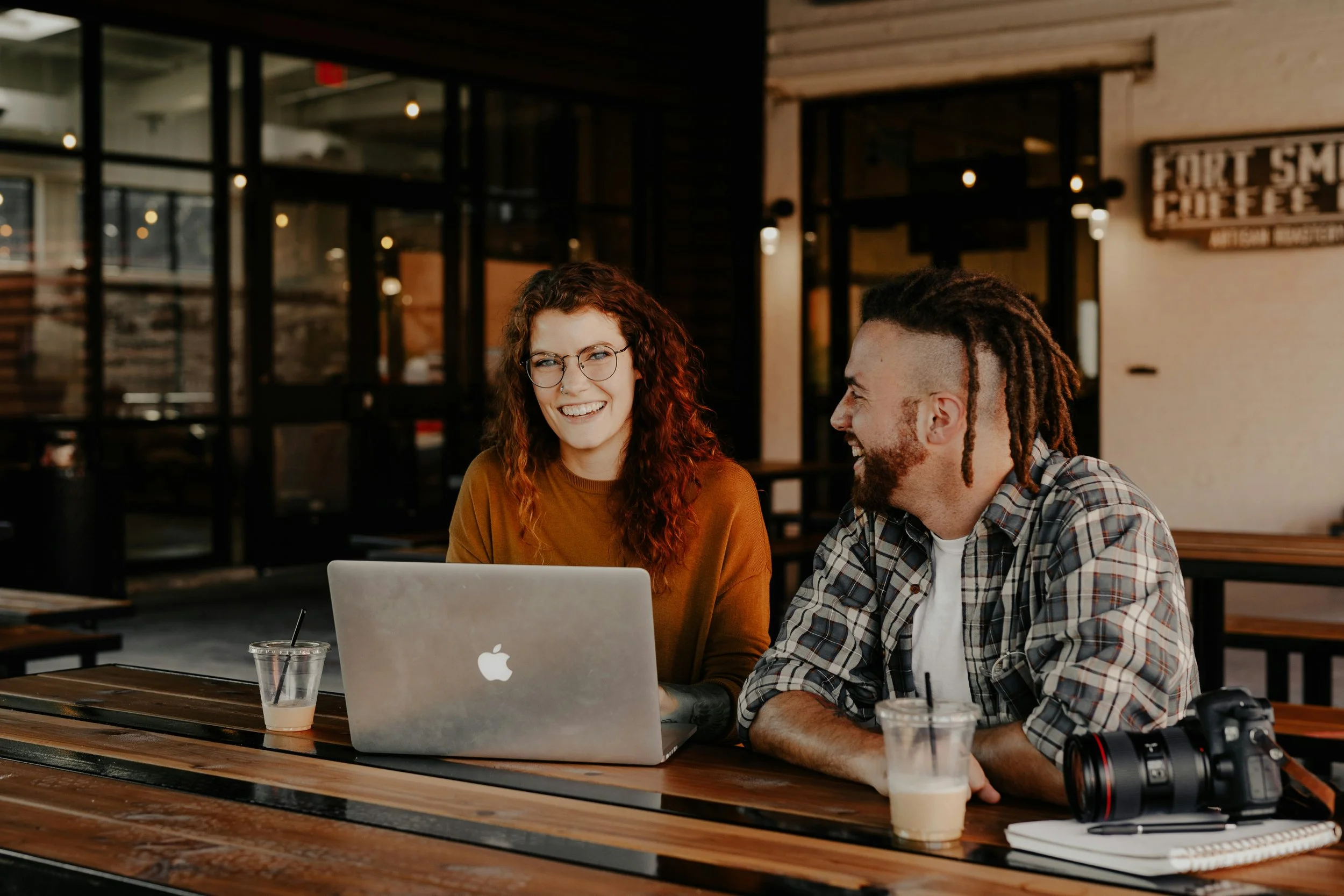 Two people, a woman with curly red hair and a man with dreadlocks and a beard, sitting at a wooden table in a coffee shop, smiling at each other, with a laptop, camera, and drinks on the table.