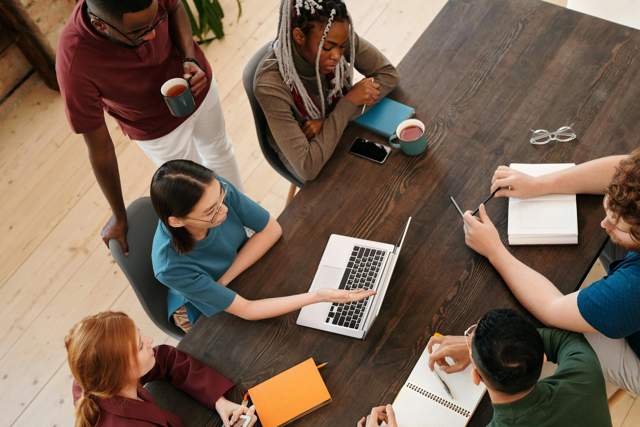 A diverse group of six people gathered around a wooden table in a meeting room. One person is sitting and pointing at a laptop screen, while others listen or take notes. Three people are standing, holding cups, and two of them are sitting, taking notes with notebooks and a pen. There are cups, a smartphone, glasses, notebooks, and a laptop on the table.