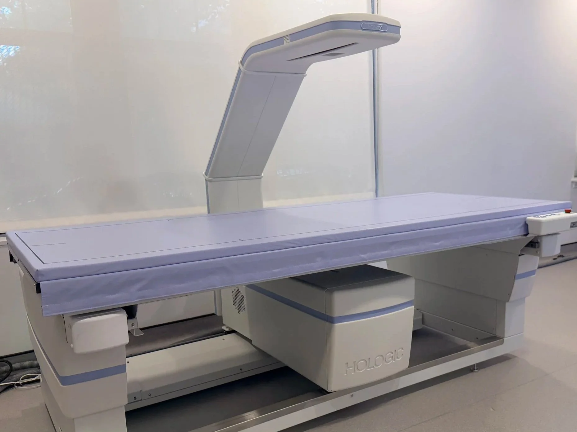A woman lying on an MRI scanning table with her eyes closed, while a technician in the background operates the machine, in a medical room with brown walls.