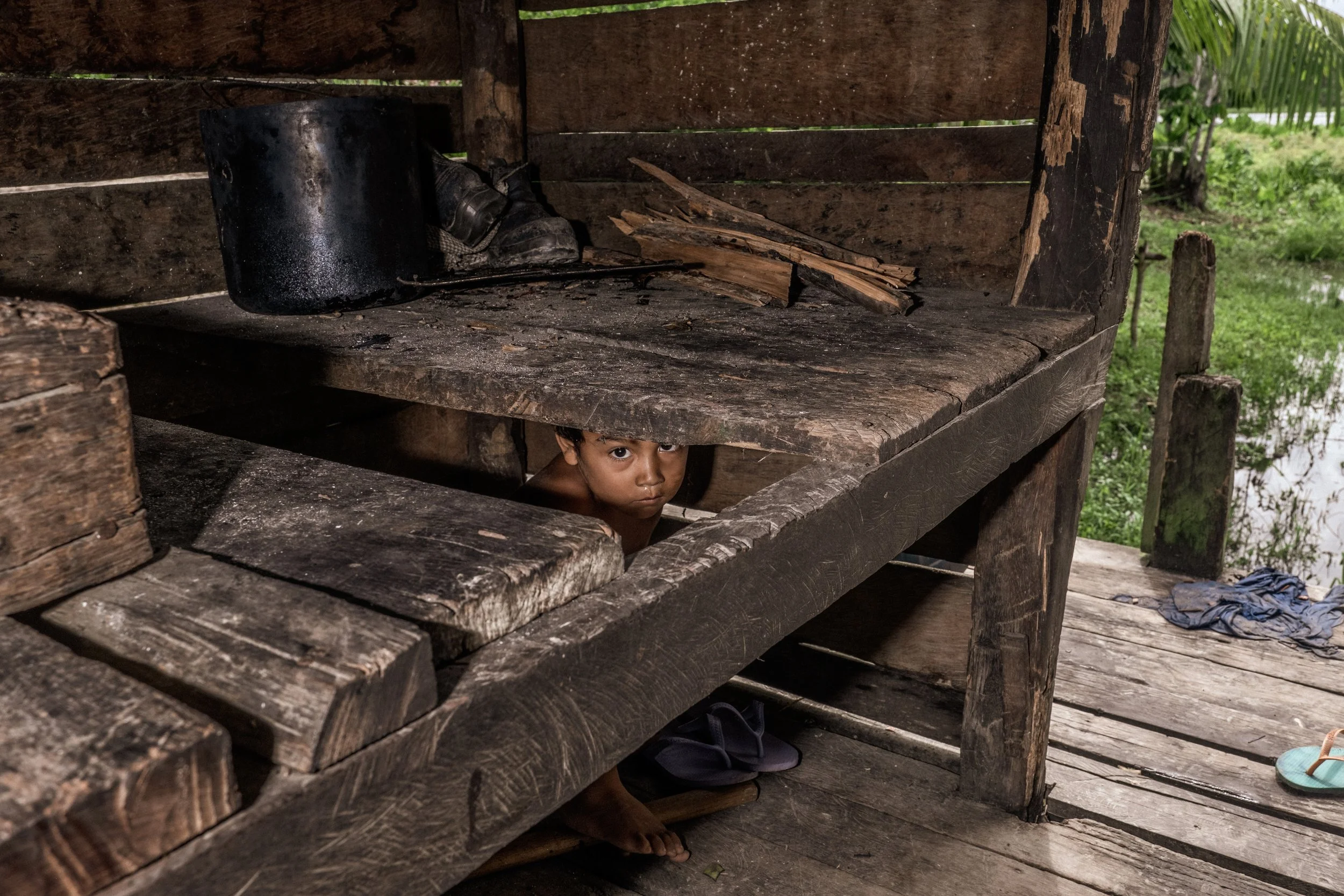Oubenson Braga, de 3 anos, se esconde sob uma estrutura de madeira usada pela família para cozinhar na casa sobre palafitas em Santa Ana, Melgaço — uma comunidade ribeirinha marcada por profunda vulnerabilidade econômica. Sem acesso à energia elétric