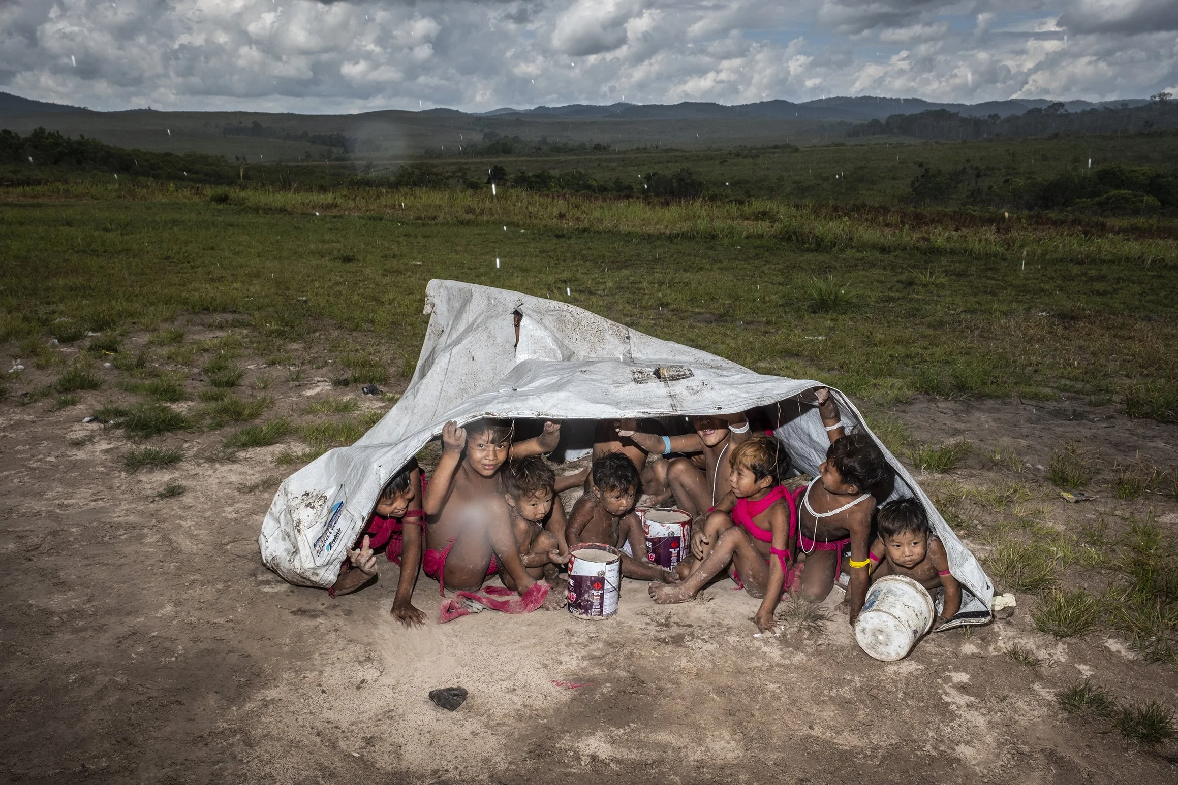 Crianças Yanomami brincam na chuva, abrigando-se sob uma lona plástica na comunidade de Surucucu.