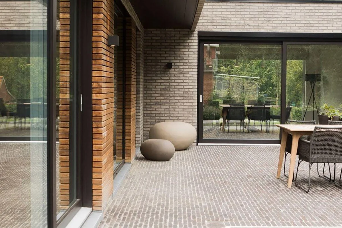 Modern outdoor patio with brick walls, glass sliding doors, a table with chairs, and decorative stone sculptures.