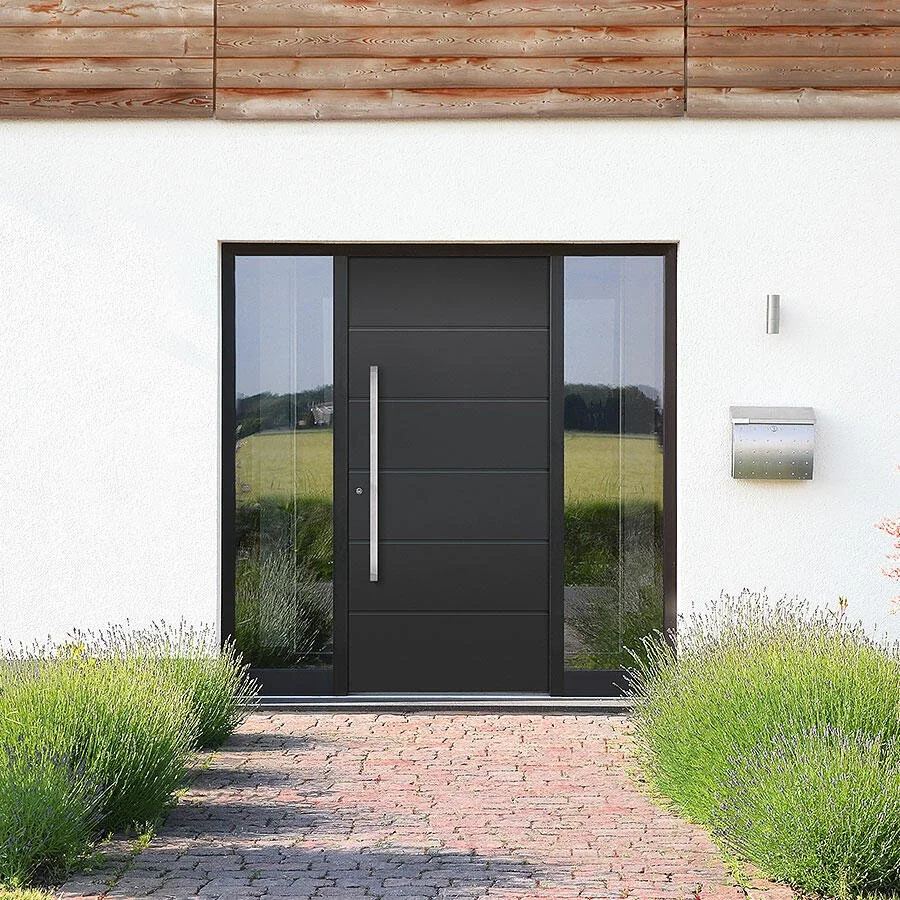 Modern front door with black panels, side glass windows, and a large vertical handle, flanked by a white wall, mailbox, and outdoor wall light, with bushes and a brick pathway leading to the entrance.