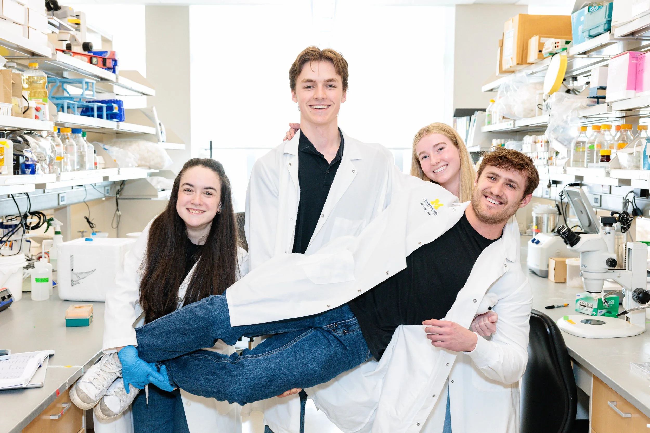 Group of five young scientists in a laboratory, smiling and holding a person lying horizontally across their arms.