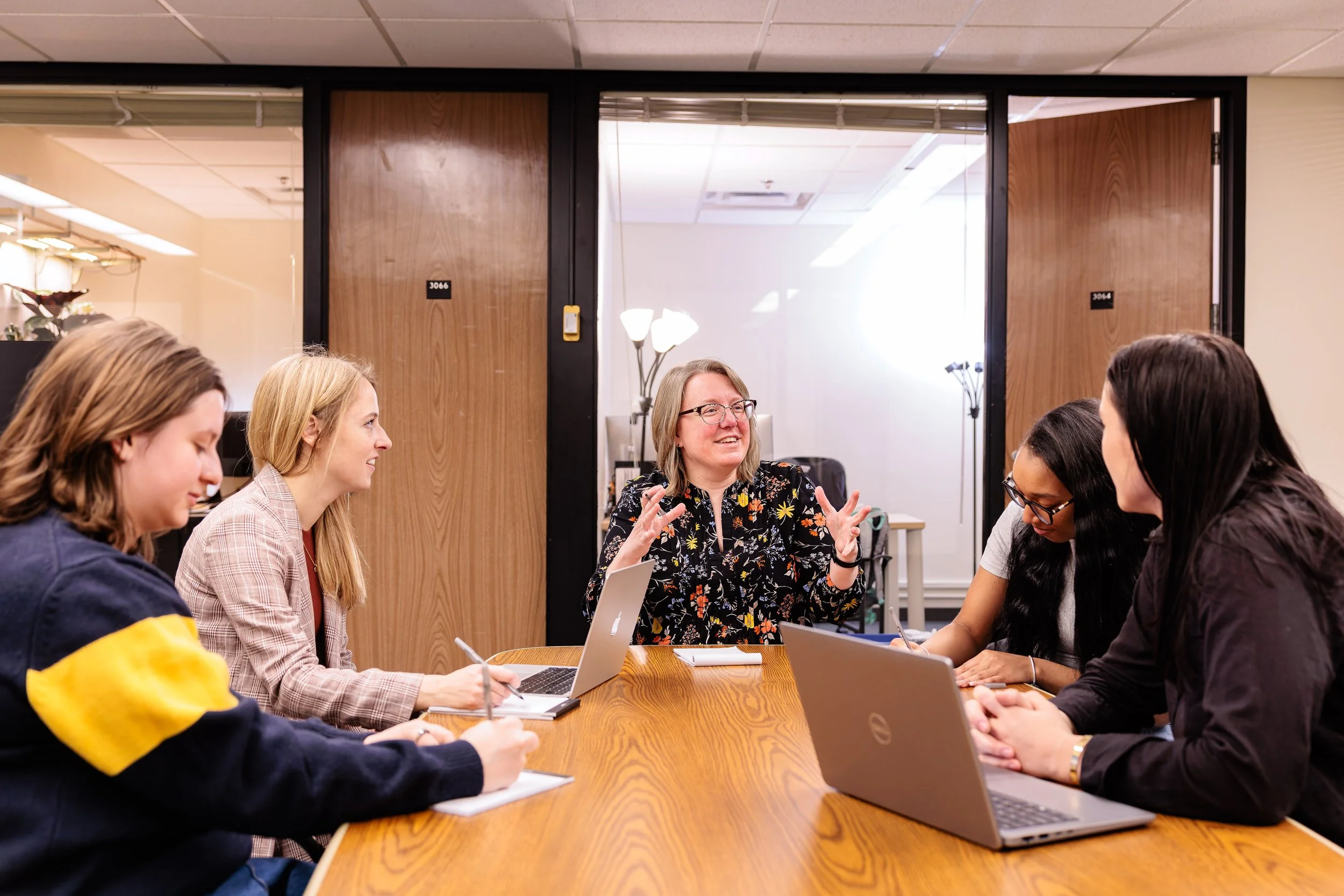 Six women in a meeting room having a discussion, some with laptops and notebooks, one woman speaking and gesturing.
