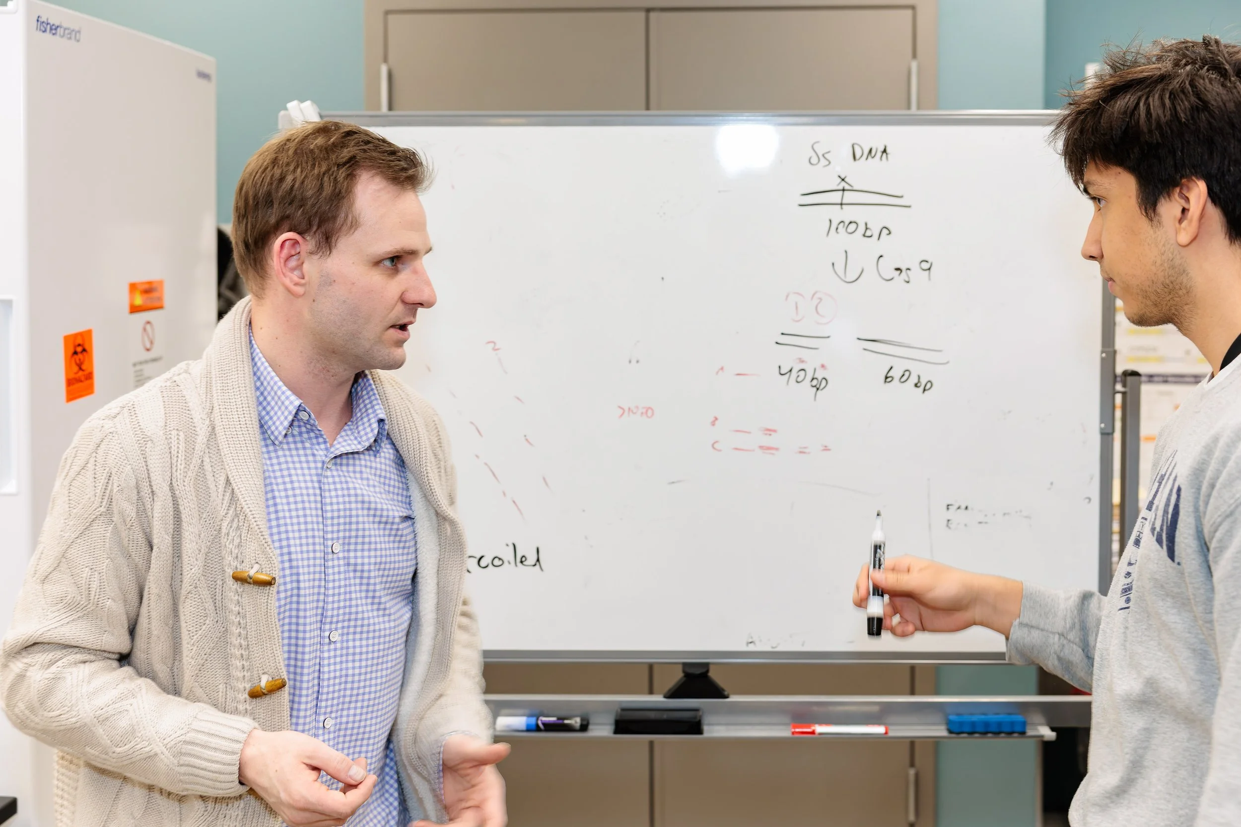 Two men having a discussion in front of a whiteboard with handwritten notes and diagrams in an office setting.