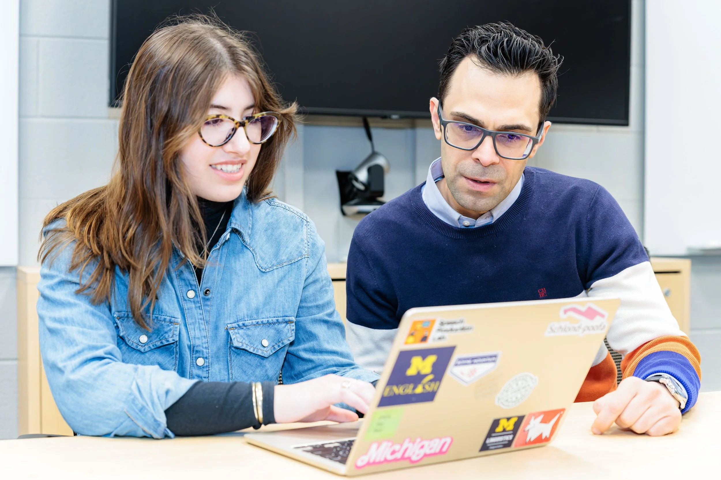 Two people, a woman and a man, sitting at a desk looking at a laptop with stickers, inside a room with a large screen or TV behind them.