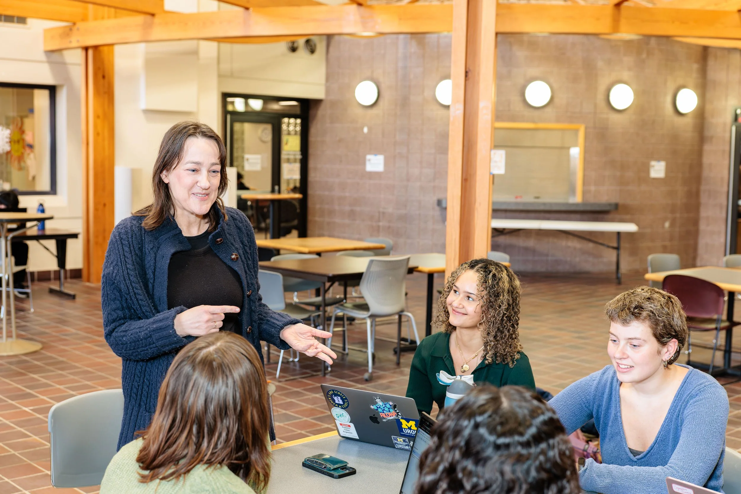 A woman standing and speaking to a group of students sitting at a table in a cafeteria or study area.