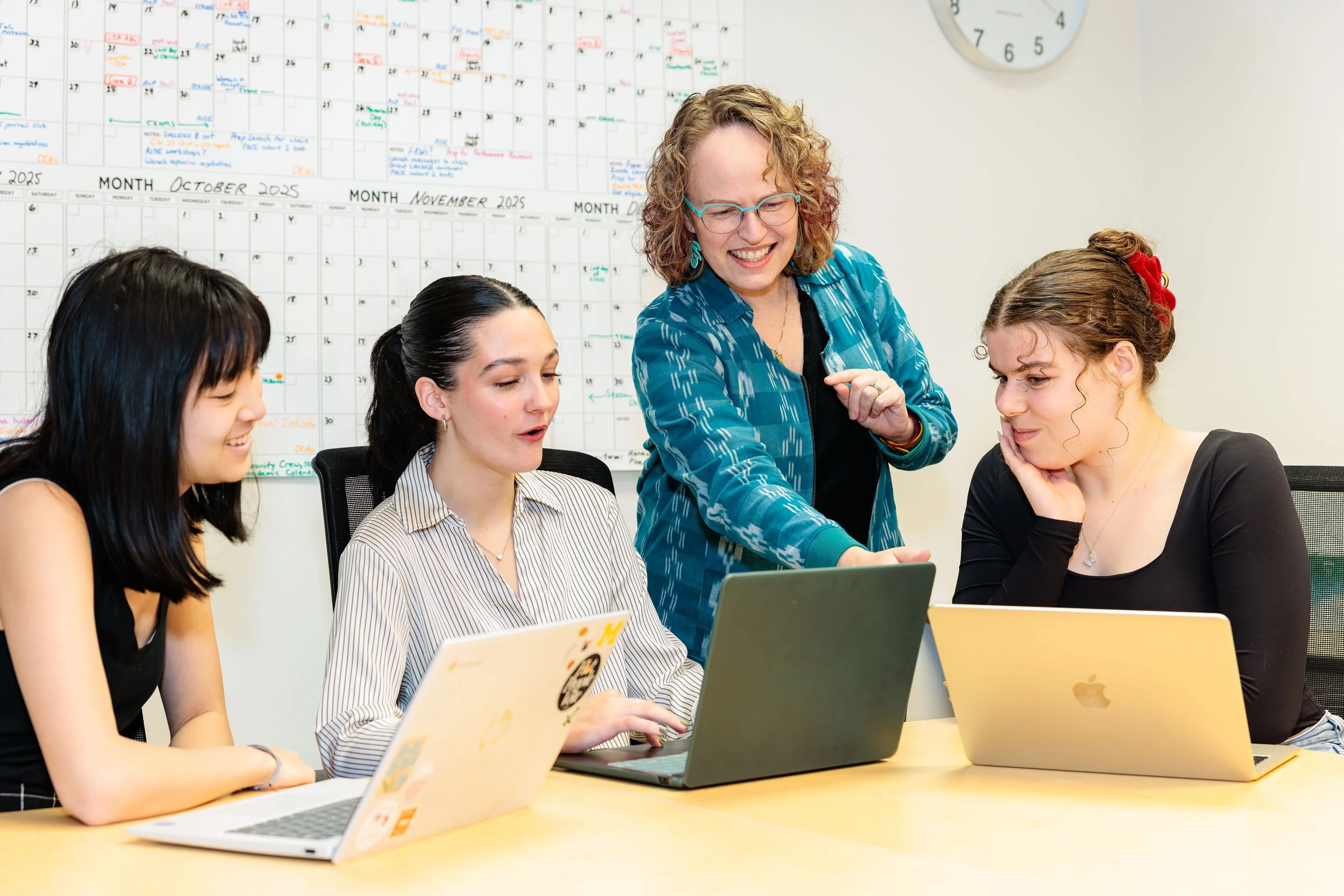 Four women gathered around a conference table with laptops, engaging in discussion. One woman stands, pointing at a laptop screen, while three women sit, looking at the screen or smiling. A whiteboard calendar for October and November 2025 is visible