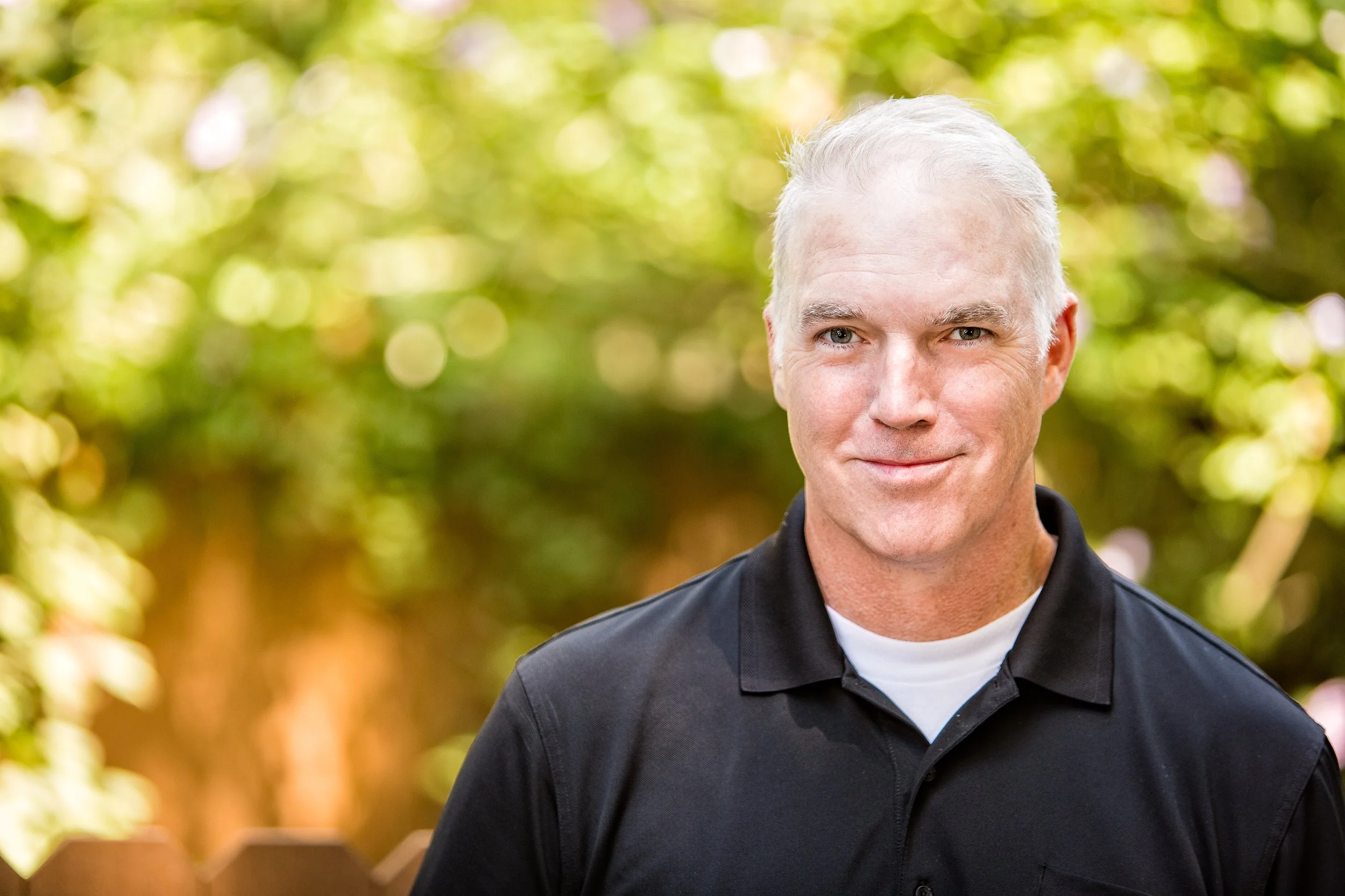 Professional headshot of a middle-aged male actor with silver hair wearing a black polo shirt against a soft outdoor background.