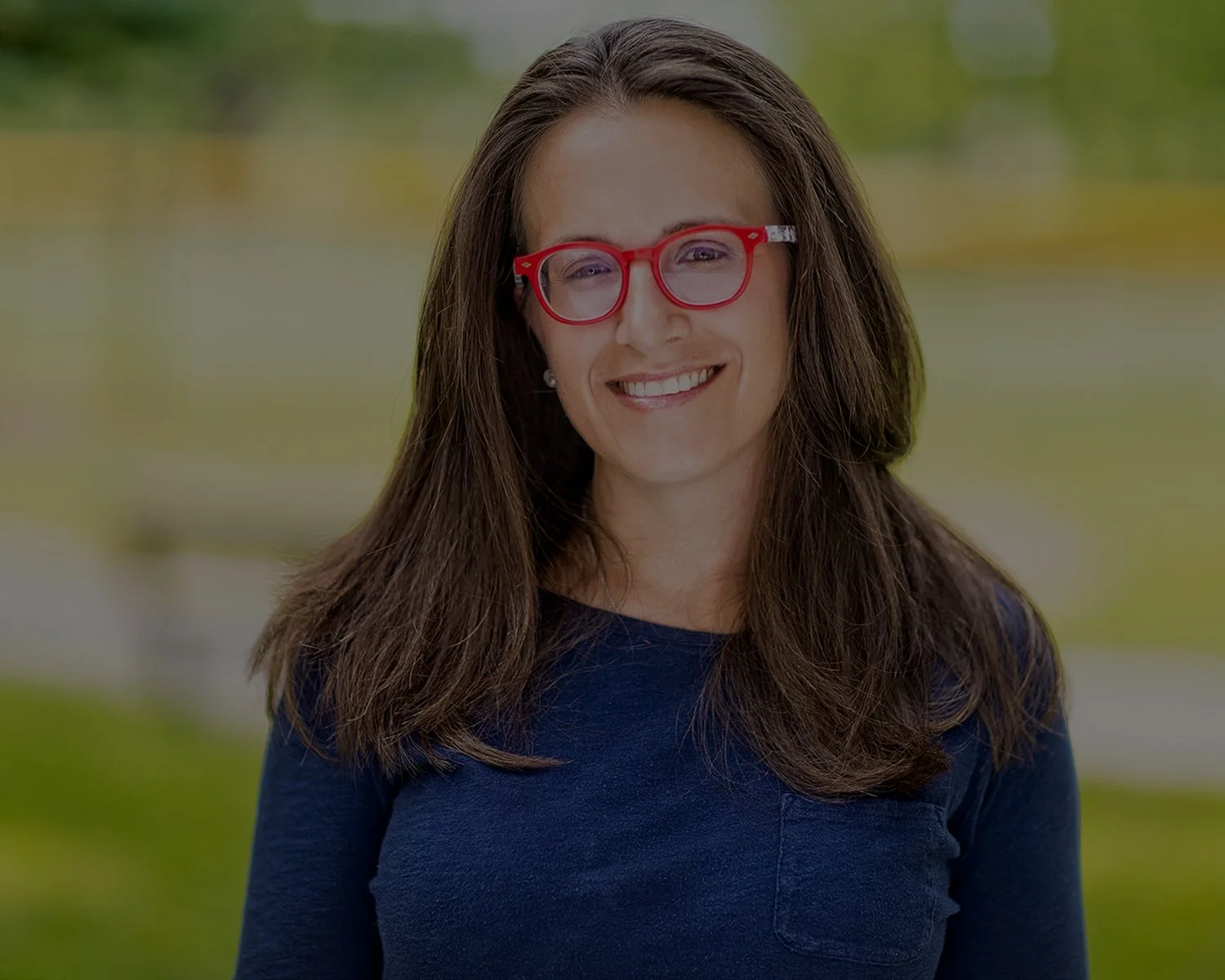 A smiling woman with long brown hair wearing red glasses and a navy blue shirt, outdoors with blurred greenery in the background.