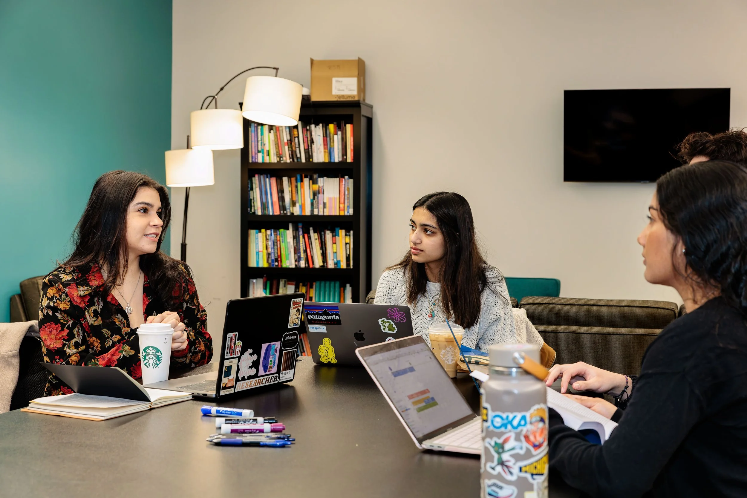 Four women sitting around a black table in a casual office, engaged in conversation, with laptops, notebooks, and drinks in front of them.