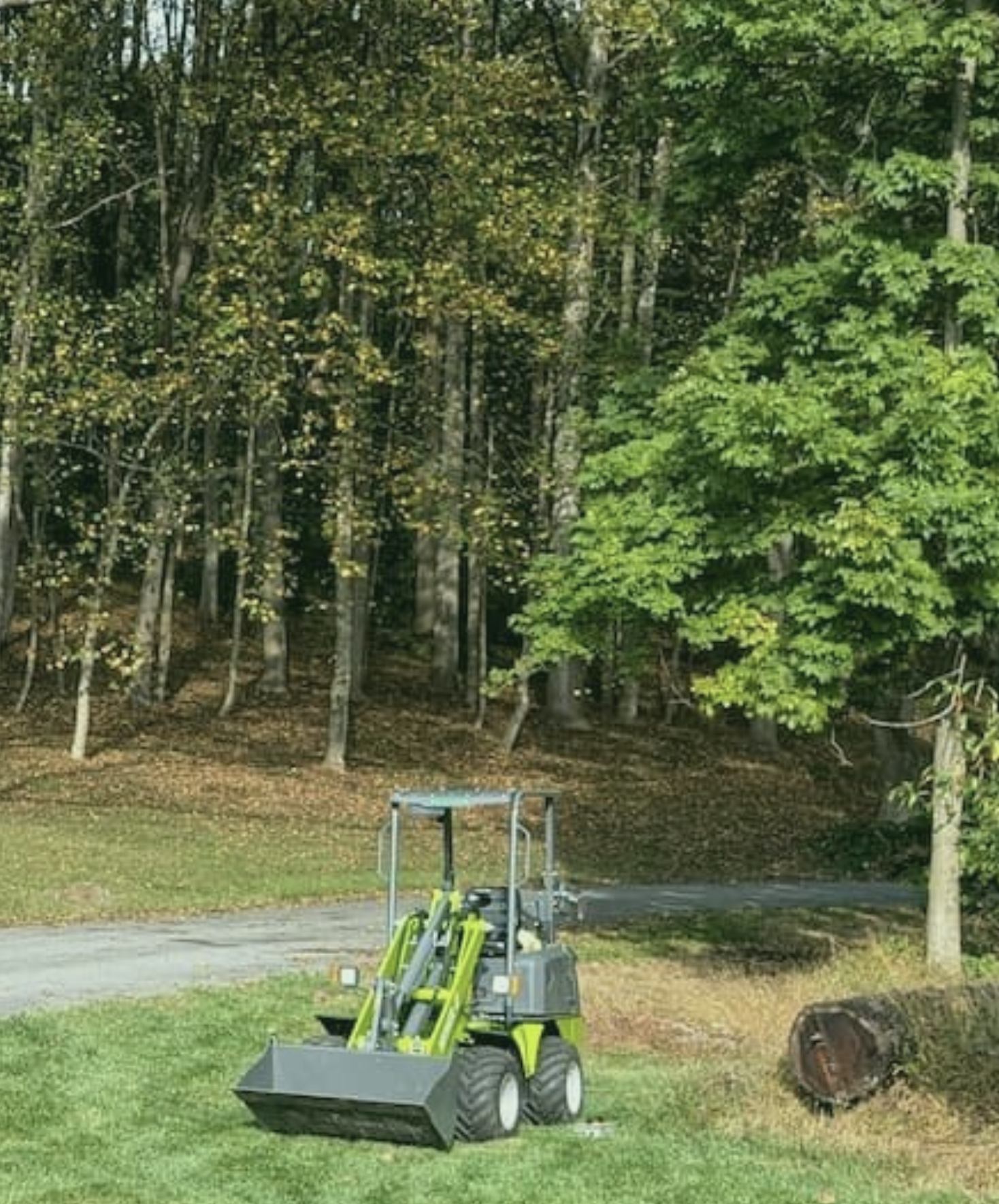A small yellow and black front loader with a bucket attachment working on a grassy lawn, near a tree trunk and a forested area.