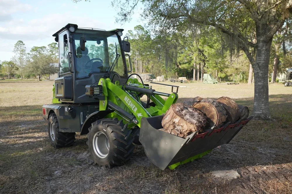 Green tractor with front loader lifting a load of cut logs in an open grassy area with trees in the background.