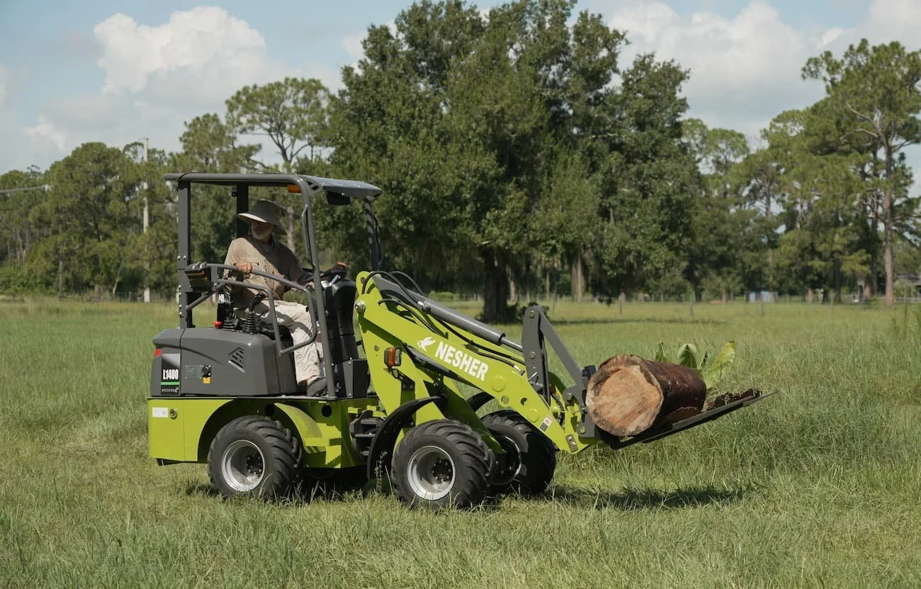 A person using a small green and gray Nesher skid steer loader to move a large log in a grassy field with trees in the background.