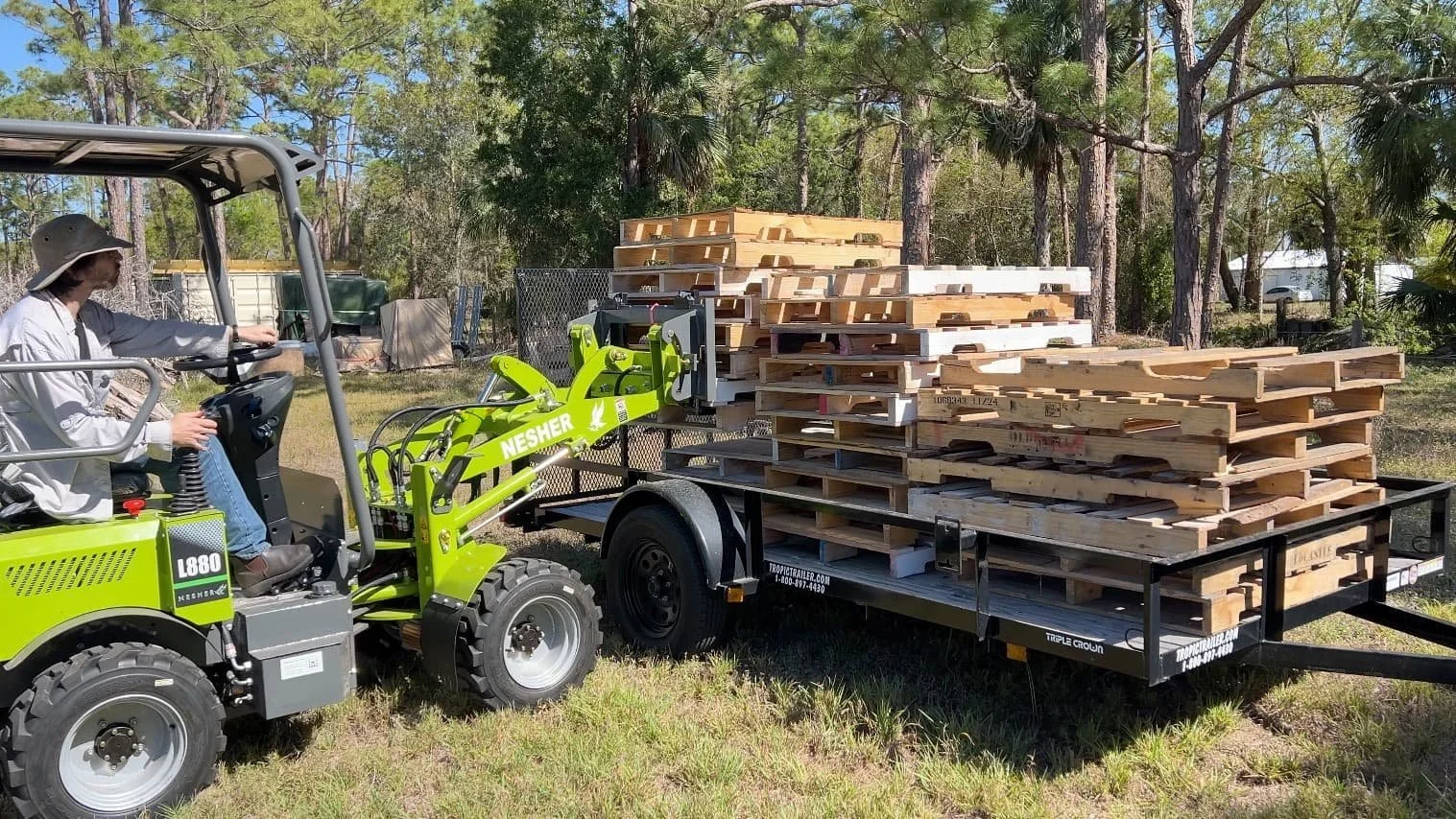 Person operating a green Nesher mini skid steer loader with pallet forks, lifting wooden pallets onto a flatbed trailer in a grassy outdoor area with trees