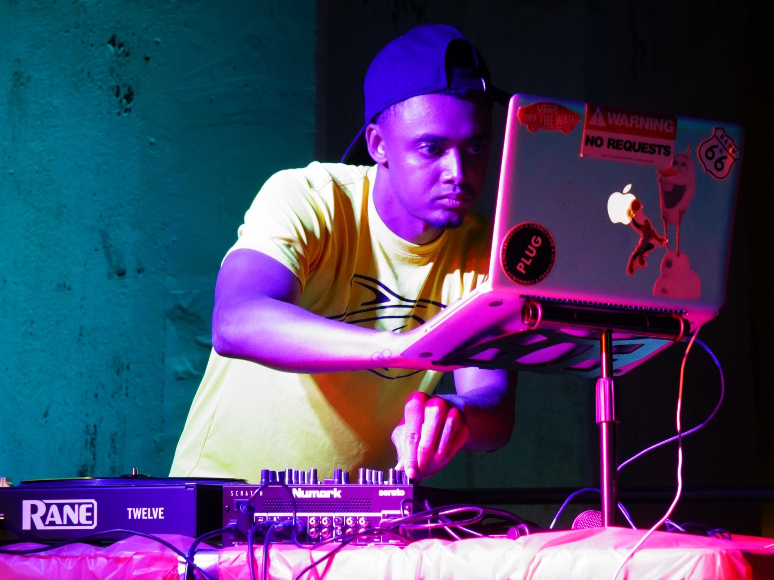 A DJ wearing a yellow T-shirt and a backward baseball cap is performing with electronic music equipment and a laptop decorated with stickers, under colorful neon lights.