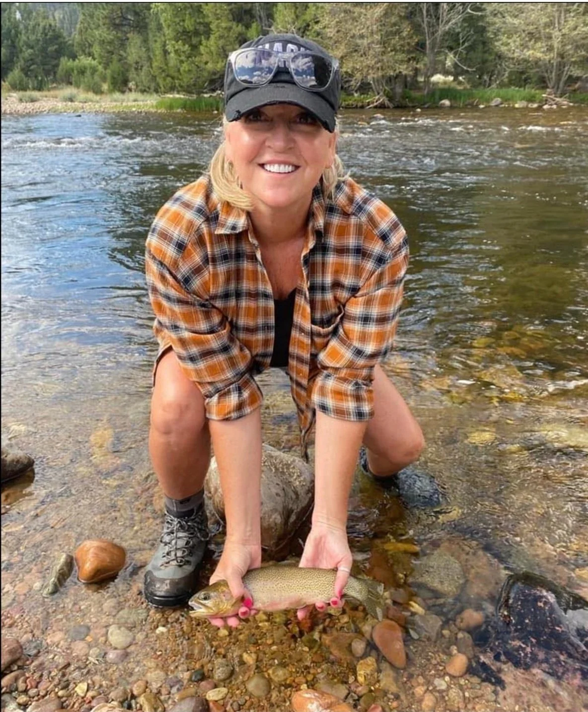 Woman crouching in a river, holding a fish she caught, wearing a plaid shirt, hat, sunglasses, and hiking boots, with trees in the background.