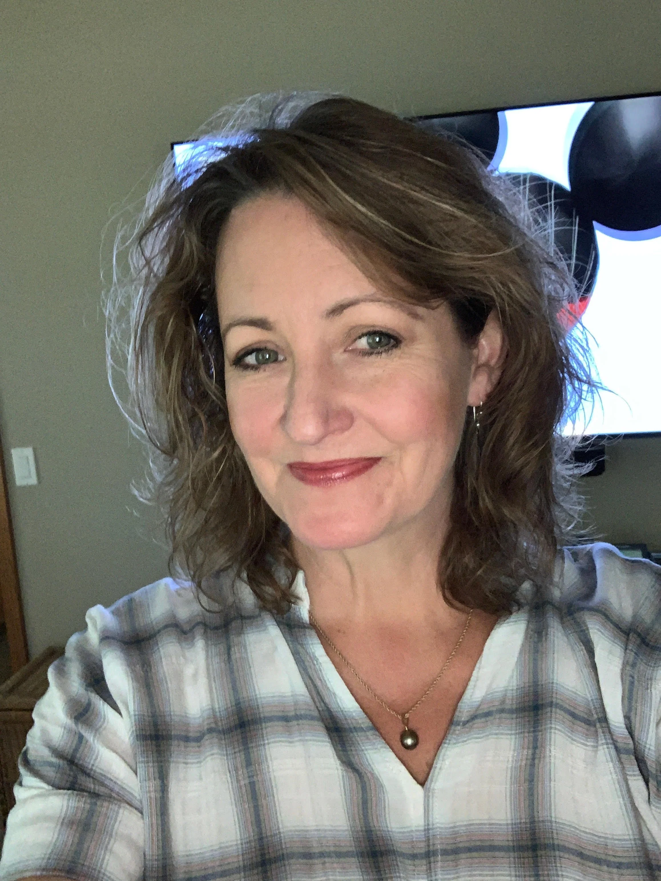 A woman with wavy brown hair, wearing a plaid shirt and a pearl necklace, smiling for a selfie.