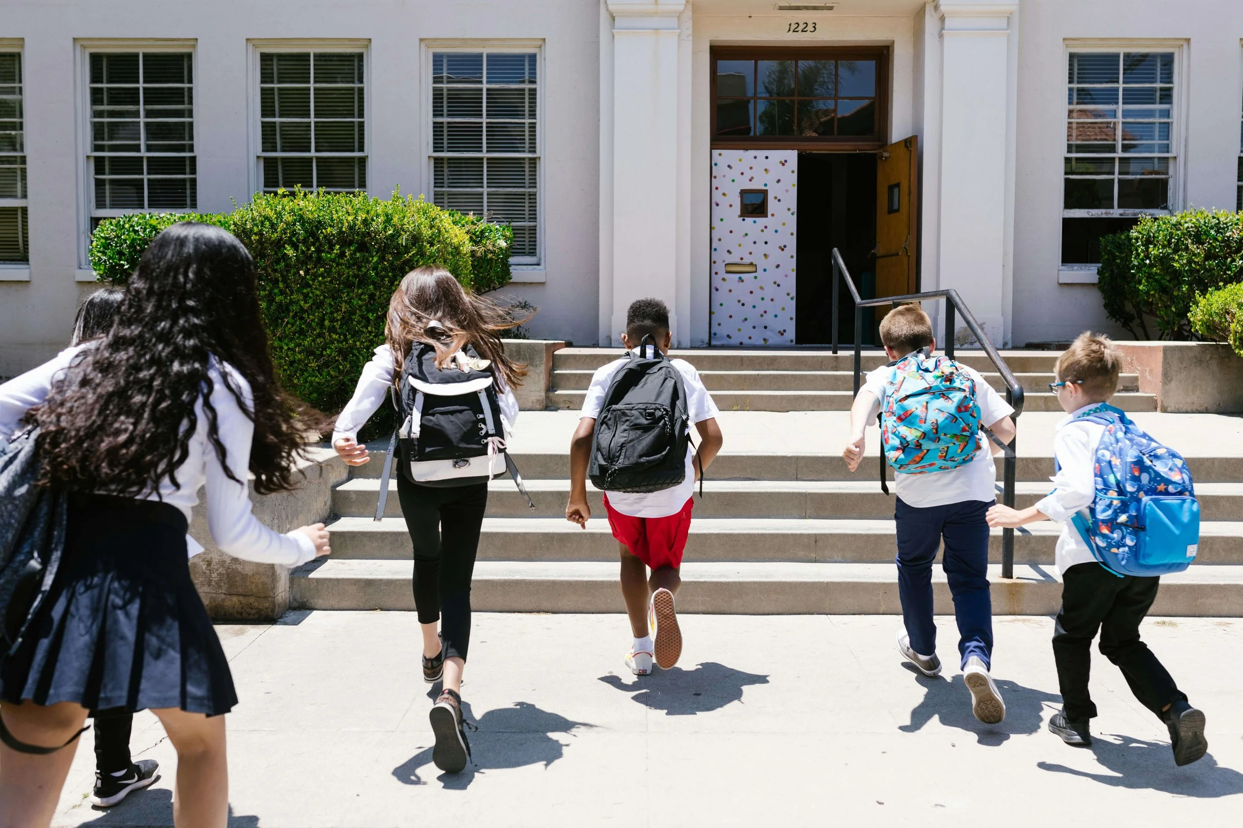 Six children with backpacks walking up stairs toward the entrance of a school building on a sunny day.