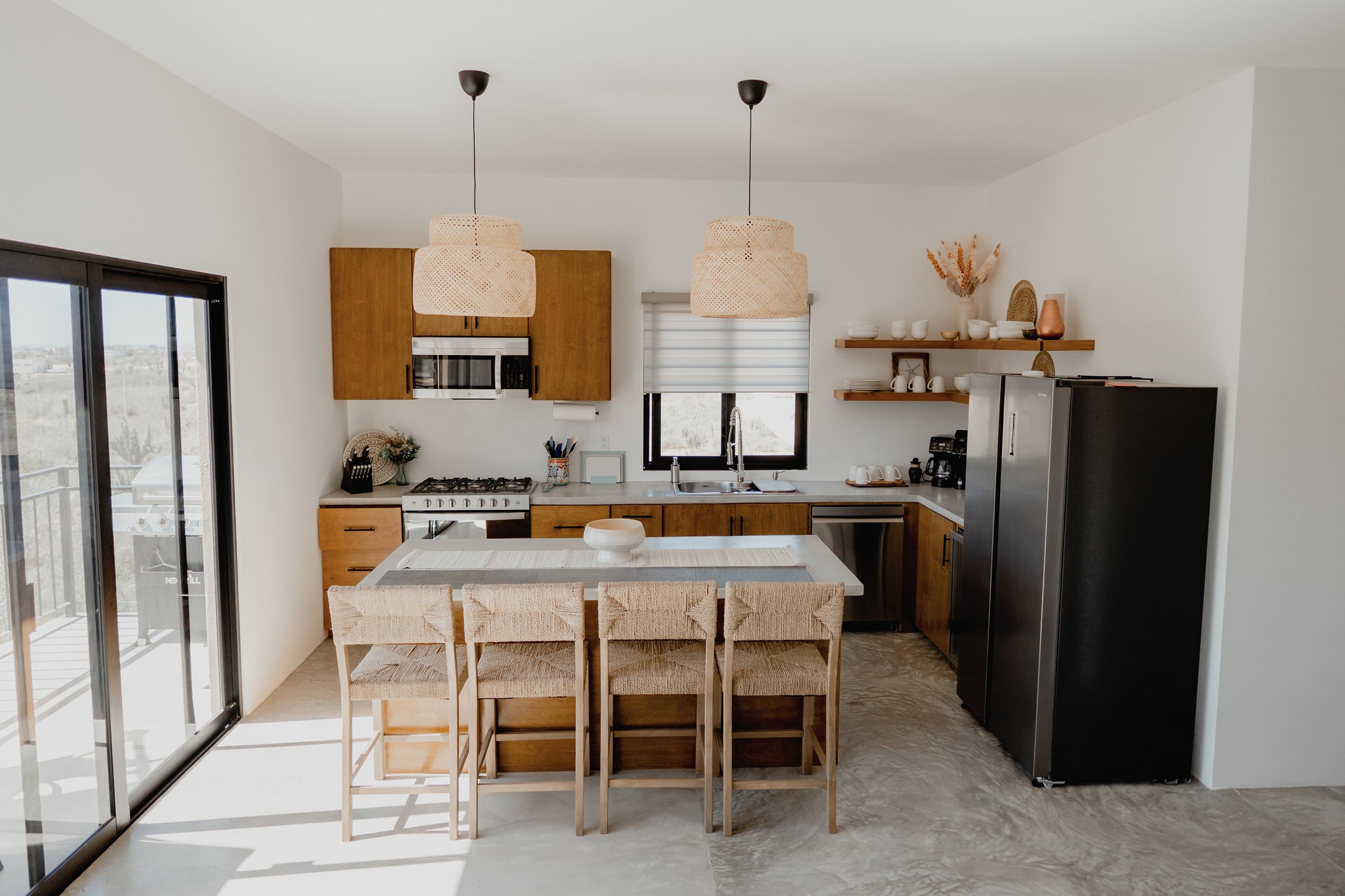 Modern kitchen with wooden cabinets, stainless steel appliances, a dining table with six woven chairs, and open shelves with dishes. Large sliding glass door lets in natural light.