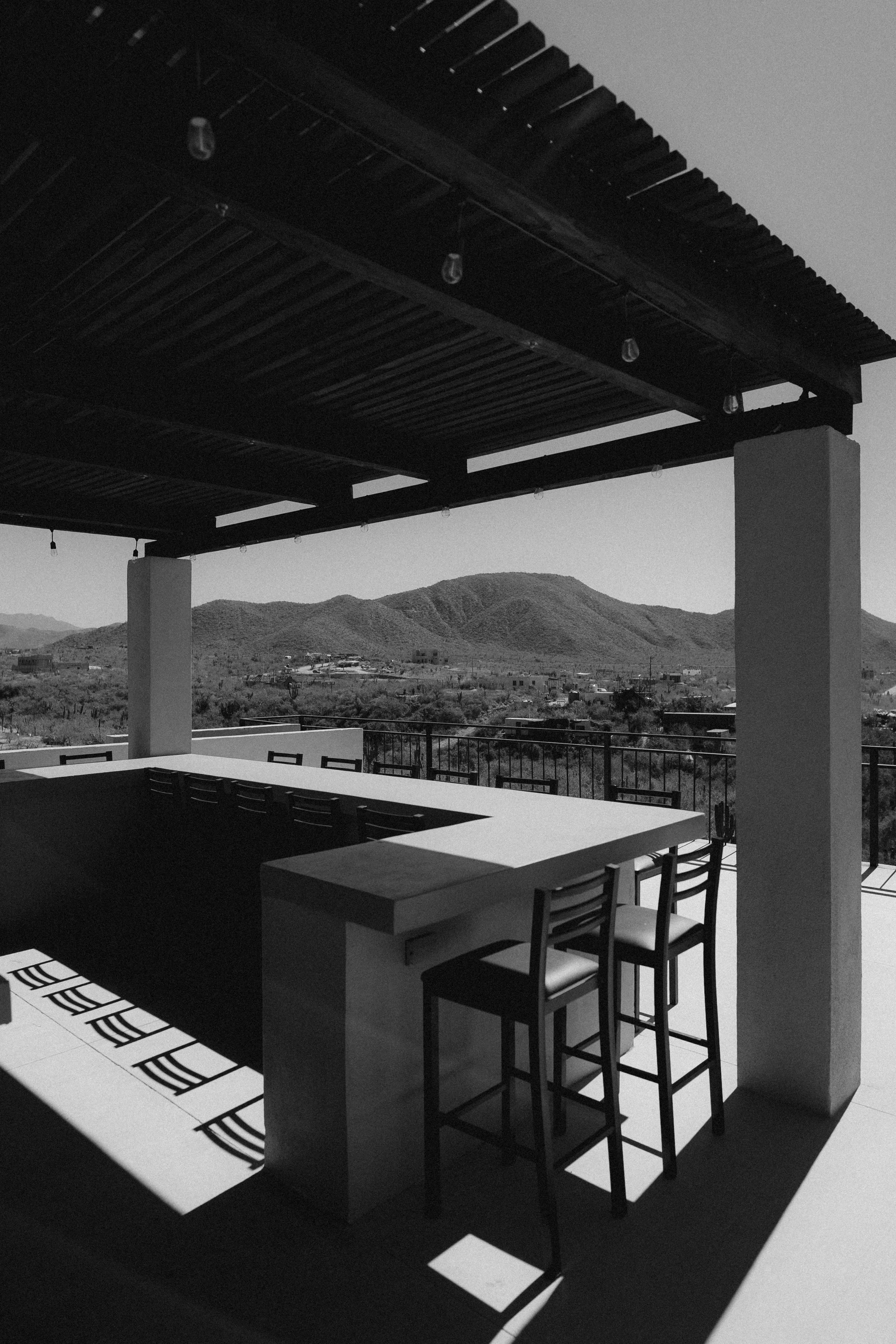 Outdoor dining area with a large table and chairs, with an overhanging roof and string lights, overlooking a mountainous landscape in the background.