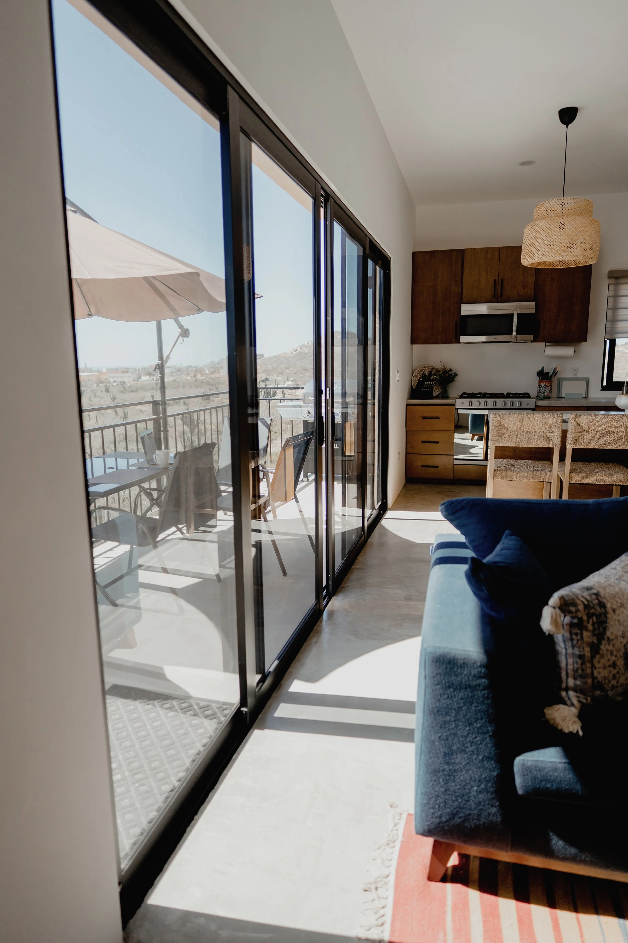 View from inside a modern apartment living room showing sliding glass doors leading to a balcony with outdoor furniture and an umbrella, overlooking a desert landscape.