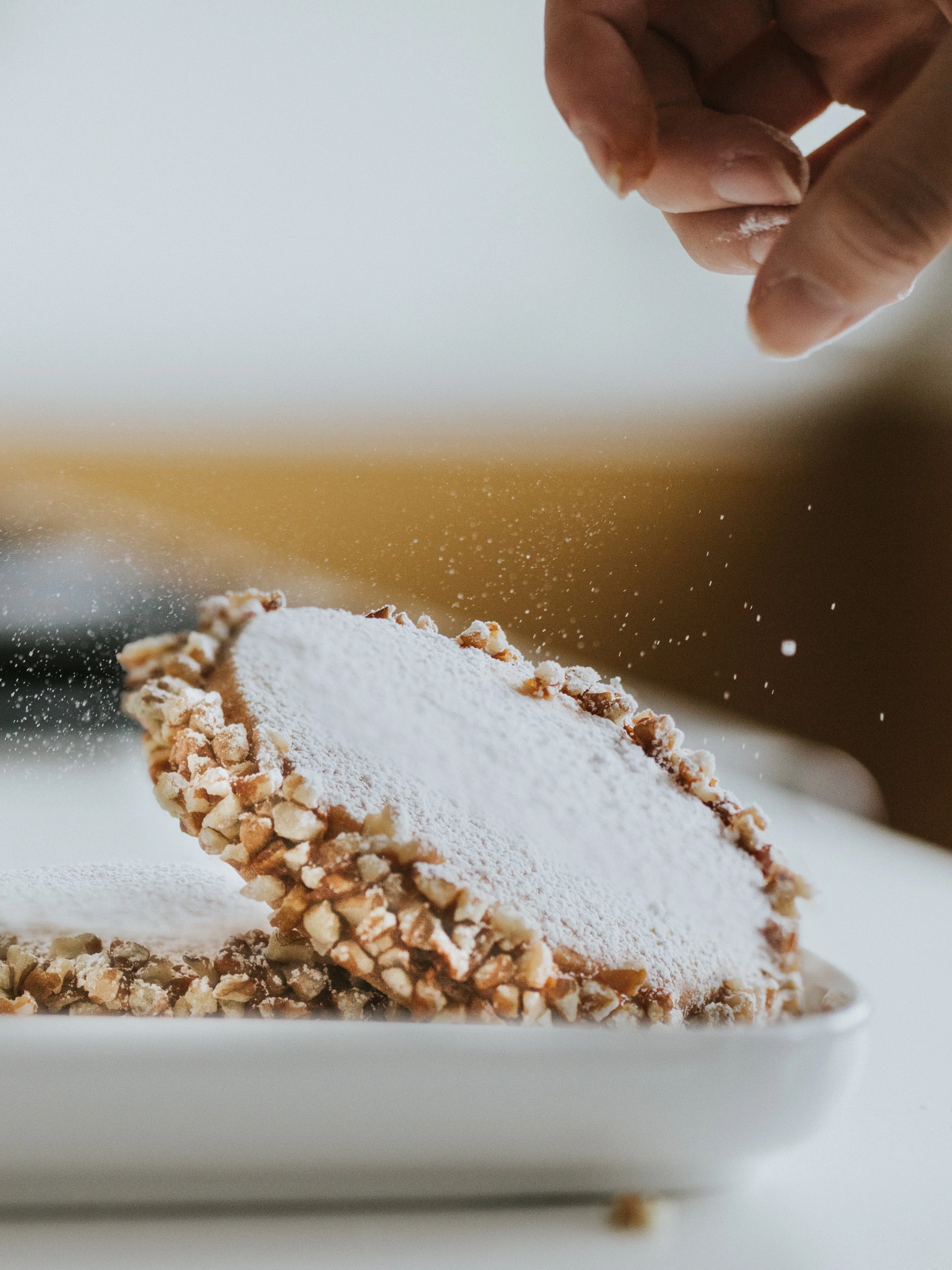 Close-up of a person sprinkling powdered sugar onto a pastry with chopped nuts on the edges.