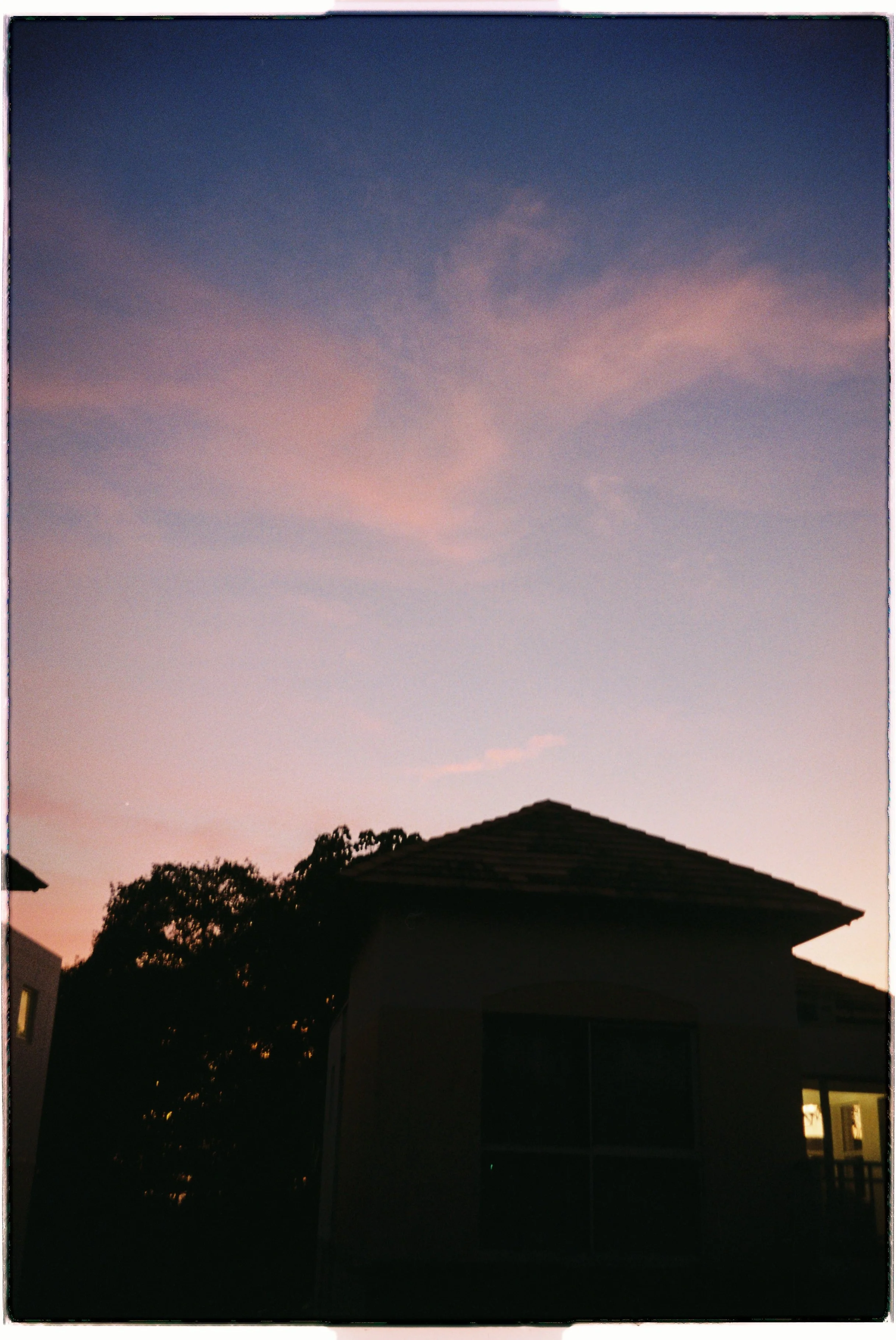 A silhouette of a house with a tiled roof, surrounded by trees, against a twilight sky with pink and purple clouds.