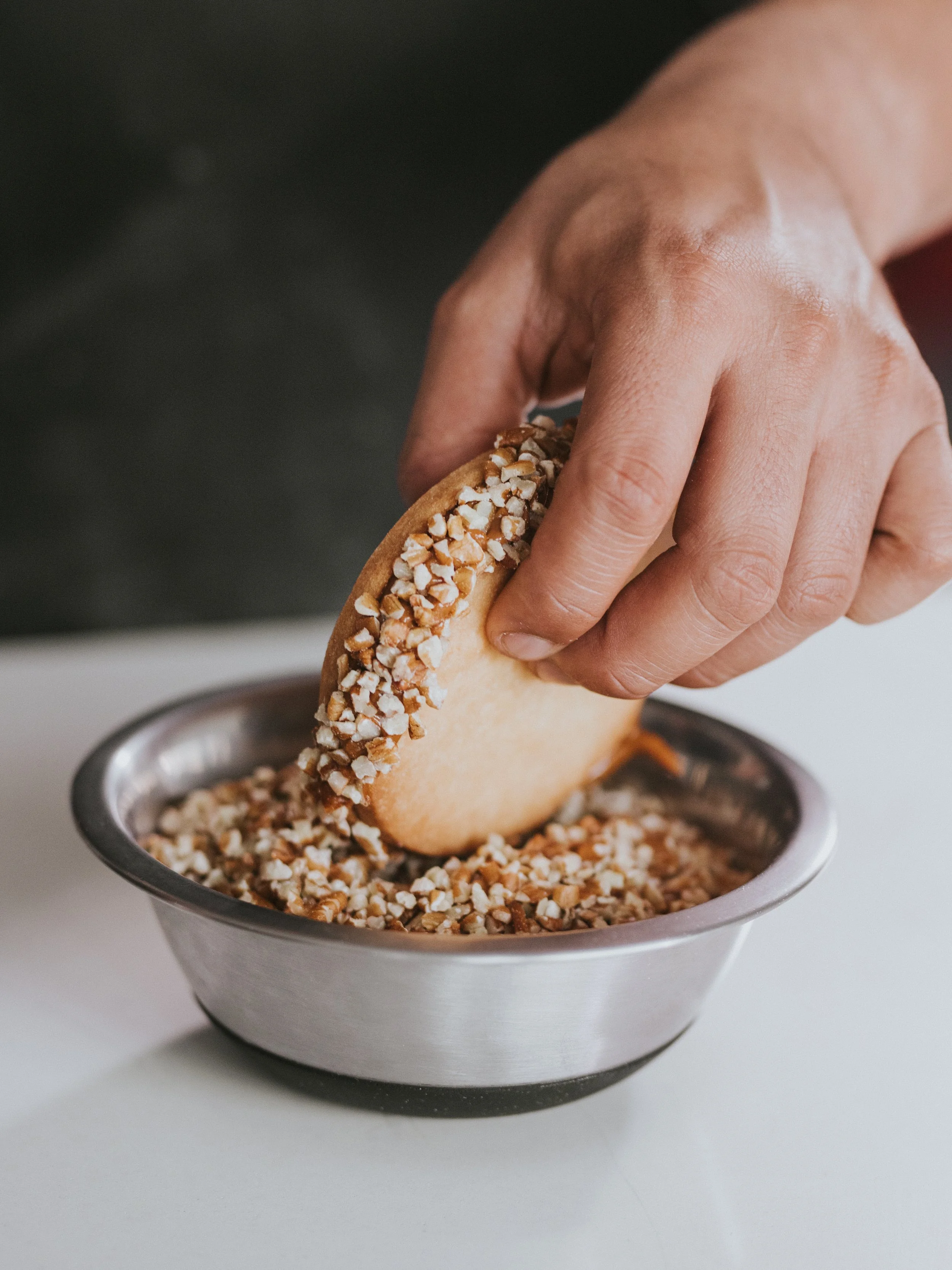 Close-up of a hand holding a cookie with chopped nuts, about to dip it into a bowl of chopped nuts.